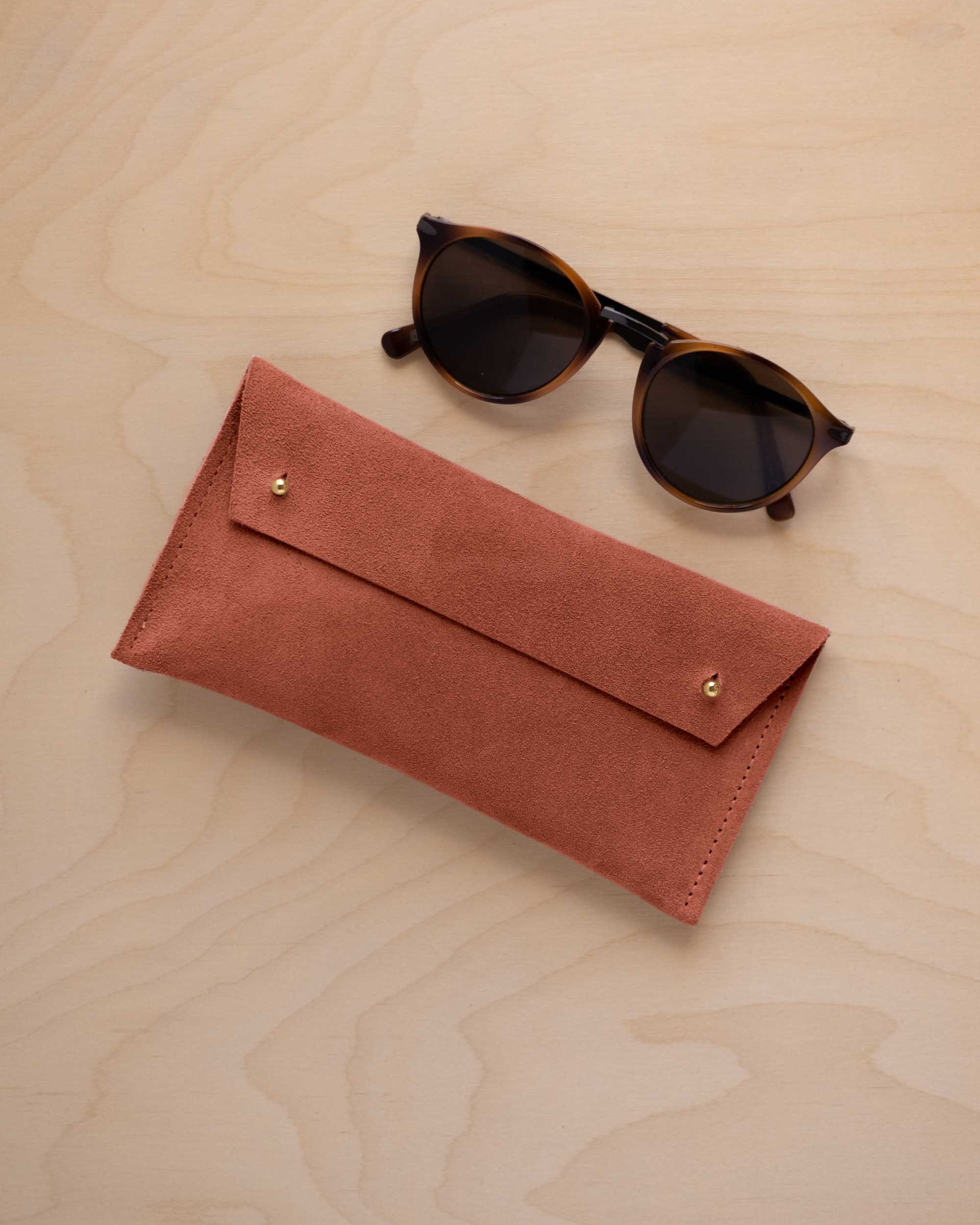 A pair of dark brown tortoiseshell sunglasses with round frames, resting on a light beige wood-grain surface next to a terracotta coloured suede glasses case with gold coloured buttons.