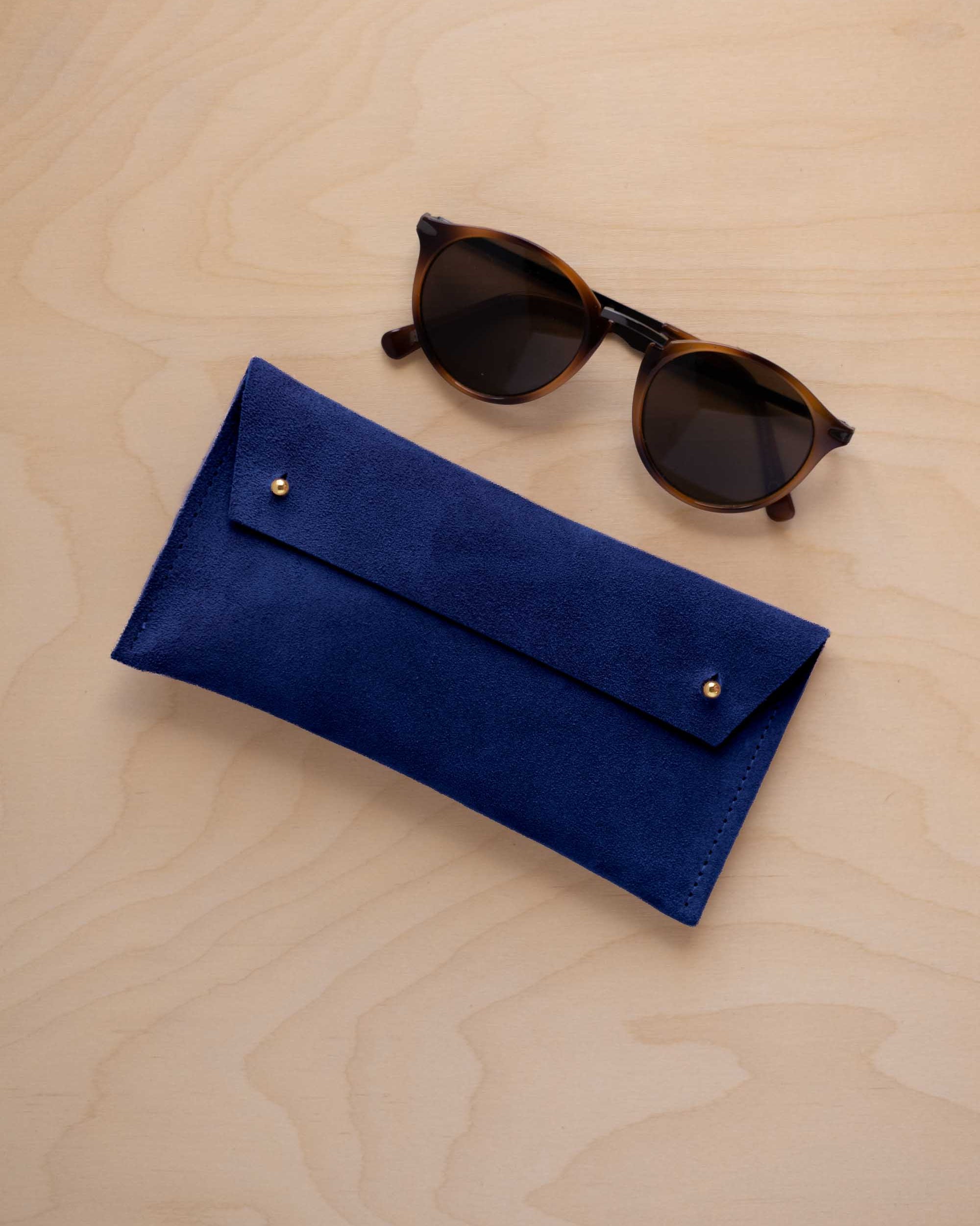 A pair of dark brown tortoiseshell sunglasses with round frames, resting on a light beige wood-grain surface next to an ultramarine blue suede glasses case with gold coloured buttons.