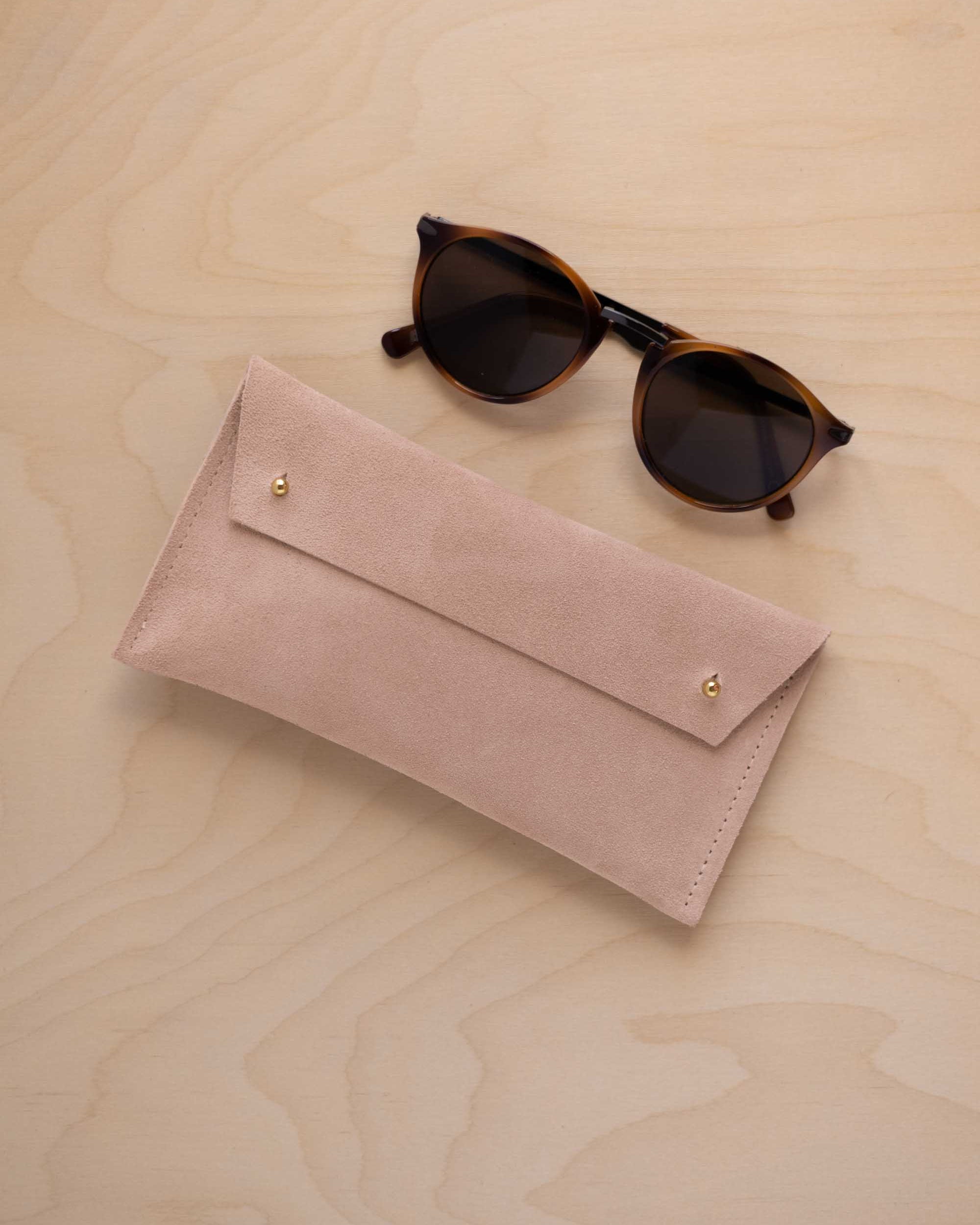 A pair of round sunglasses with dark brown tortoiseshell frames and dark lenses, resting on a light beige wood-grain surface next to a warm beige suede glasses case with gold coloured buttons.