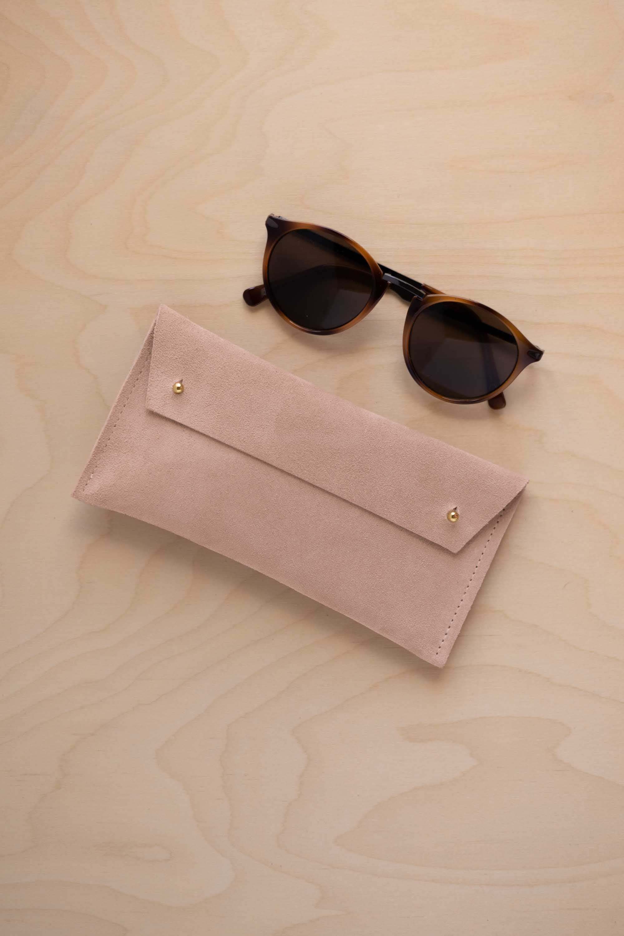 A pair of round sunglasses with dark brown tortoiseshell frames and dark lenses, resting on a light beige wood-grain surface next to a warm beige suede glasses case with gold coloured buttons.