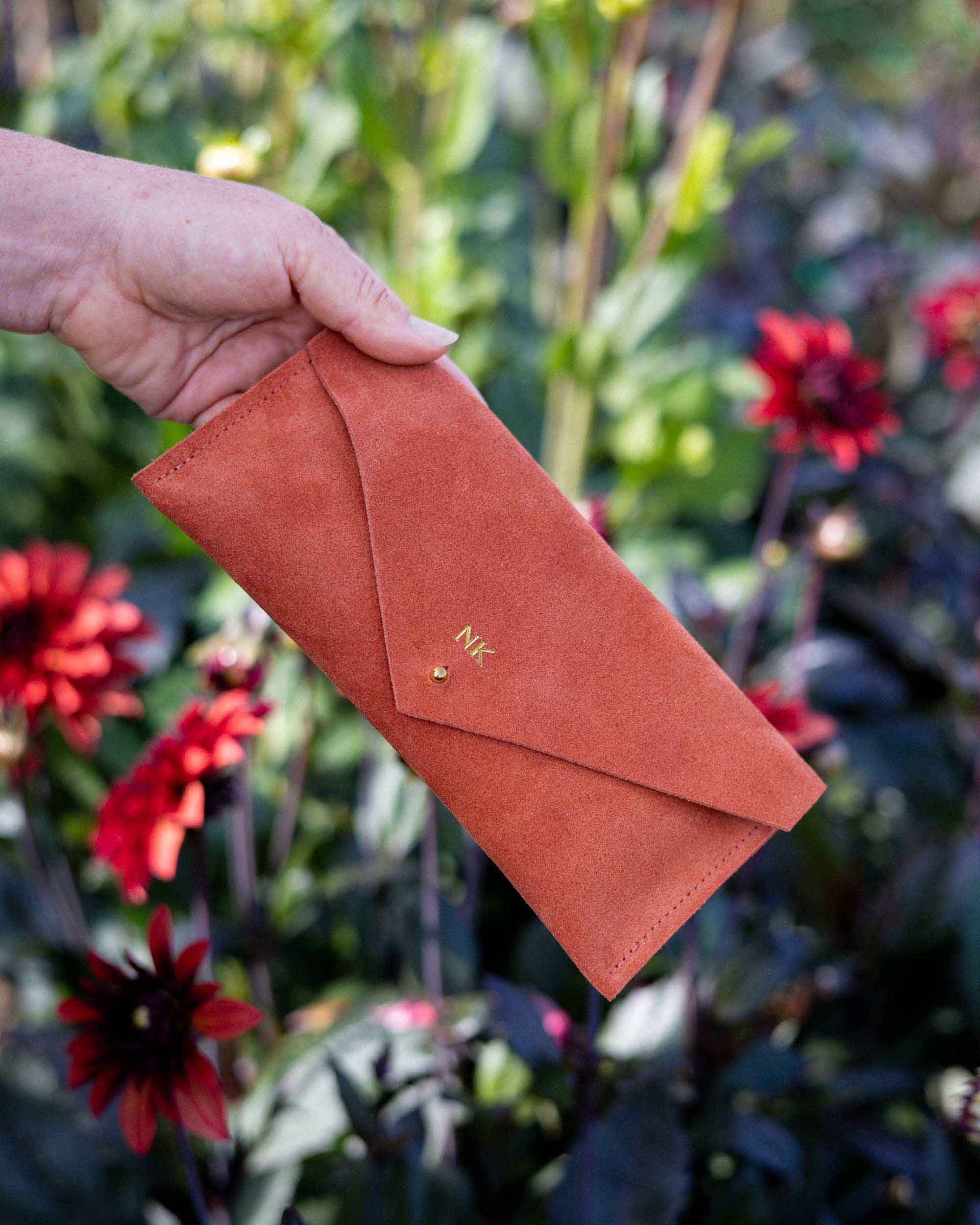 A hand holding a terracotta-colored suede envelope-style wallet or clutch with a small gold monogram, set against a blurred garden background with red dahlia flowers.