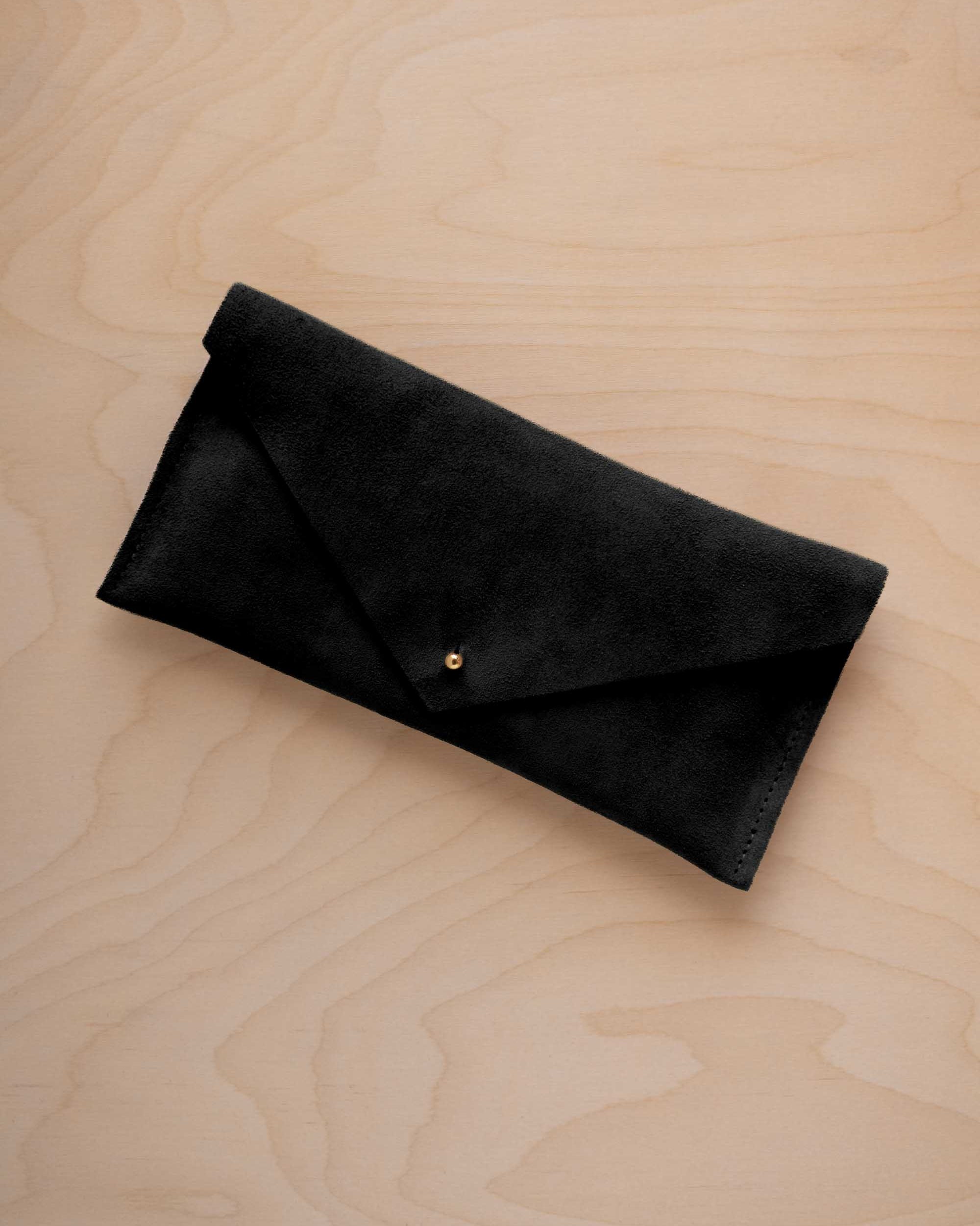 A black suede envelope clutch wallet or bag with a small gold button clasp, resting on a light beige wood grain background.