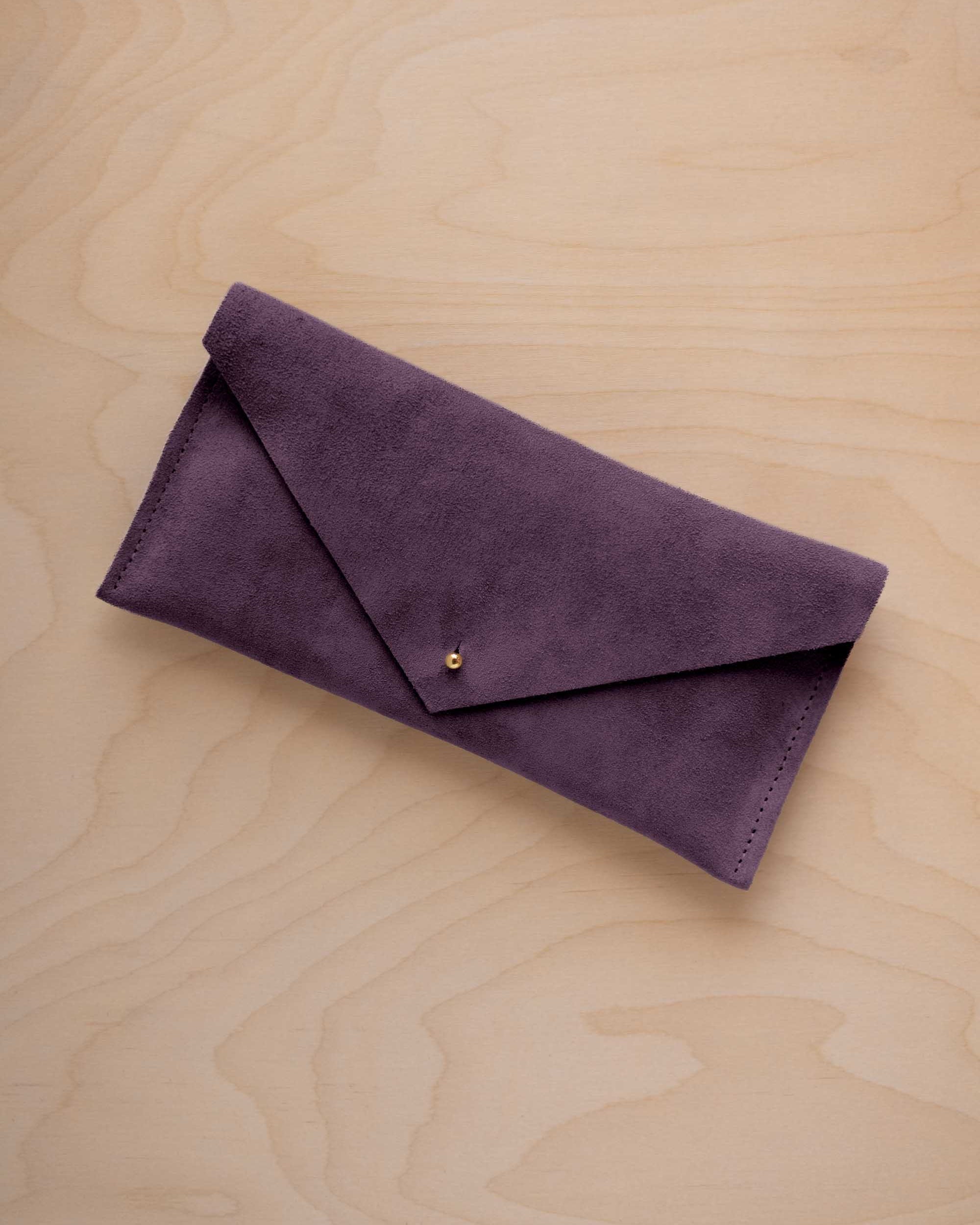 A blackcurrant dark purple coloured suede envelope clutch wallet or bag with a small gold button clasp, resting on a light beige wood grain background.