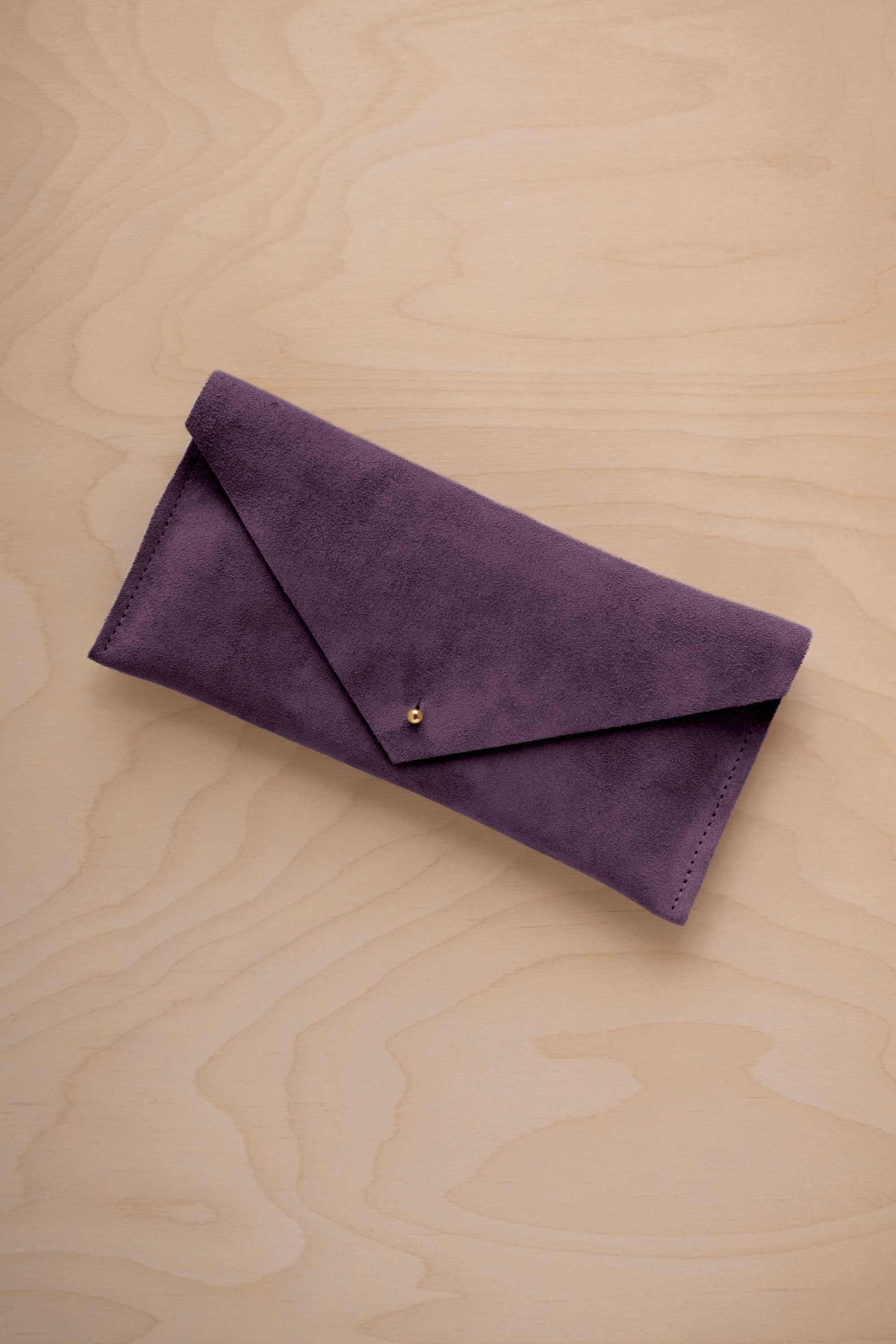 A blackcurrant dark purple coloured suede envelope clutch wallet or bag with a small gold button clasp, resting on a light beige wood grain background.