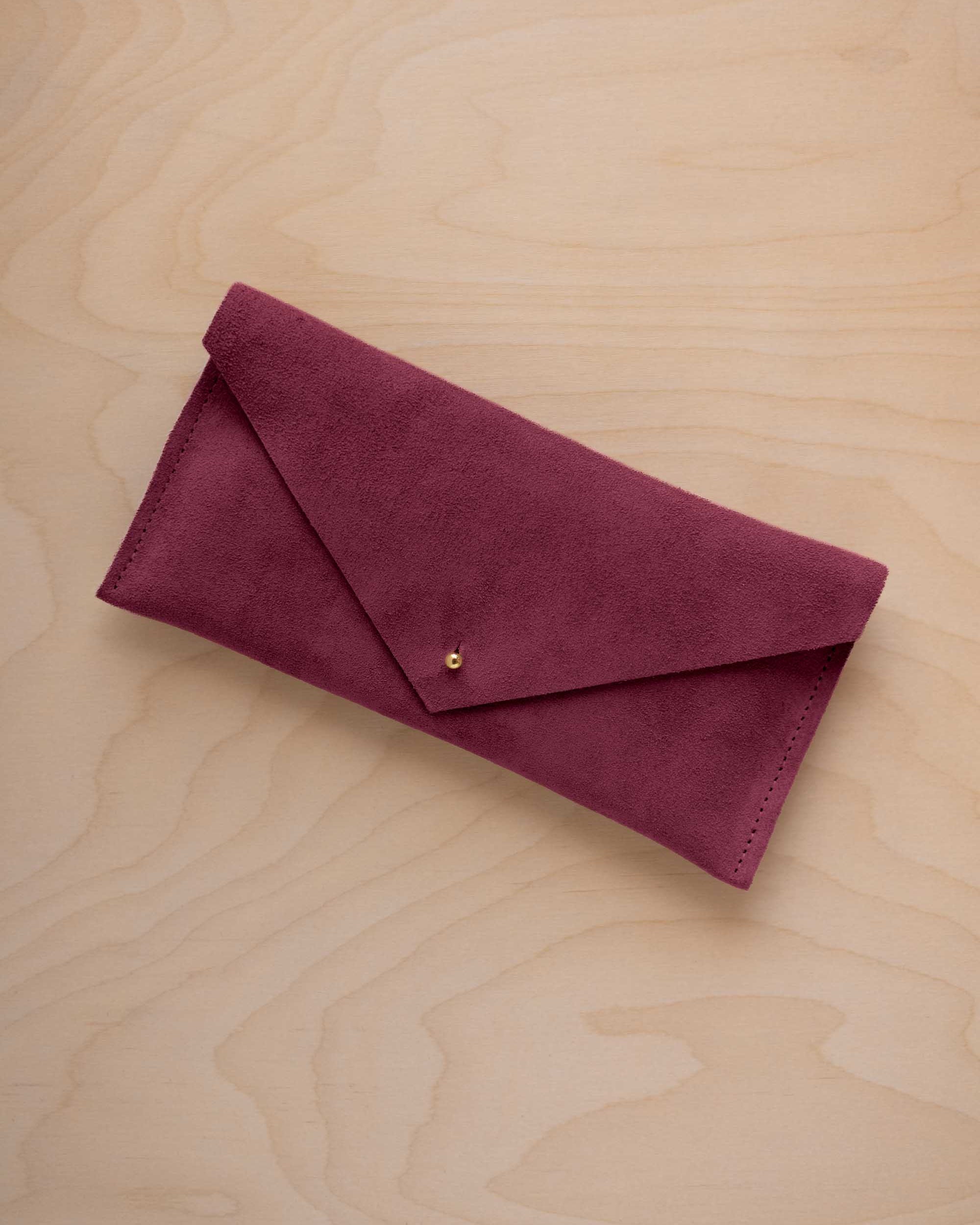 A burgundy suede envelope clutch wallet or bag with a small gold button clasp, resting on a light beige wood grain background.