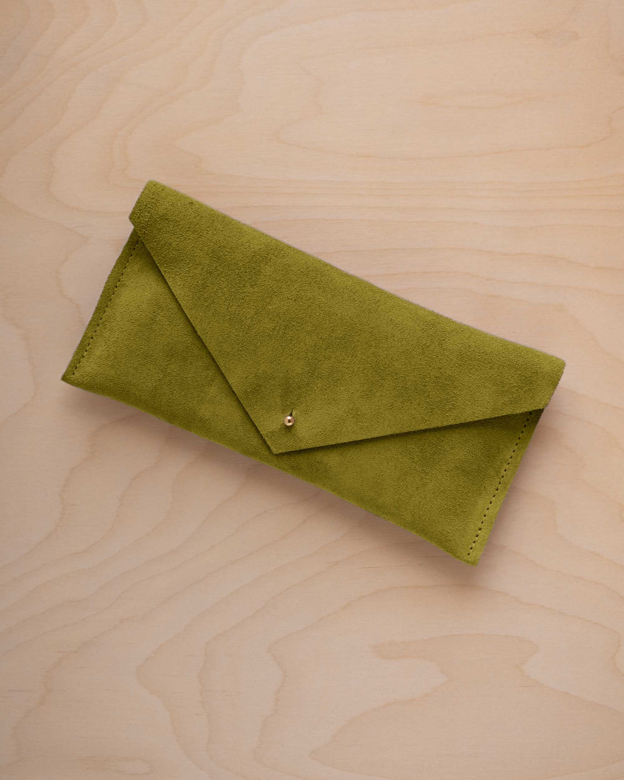 A chartreuse lime green coloured suede envelope clutch wallet or bag with a small gold button clasp, resting on a light beige wood grain background.