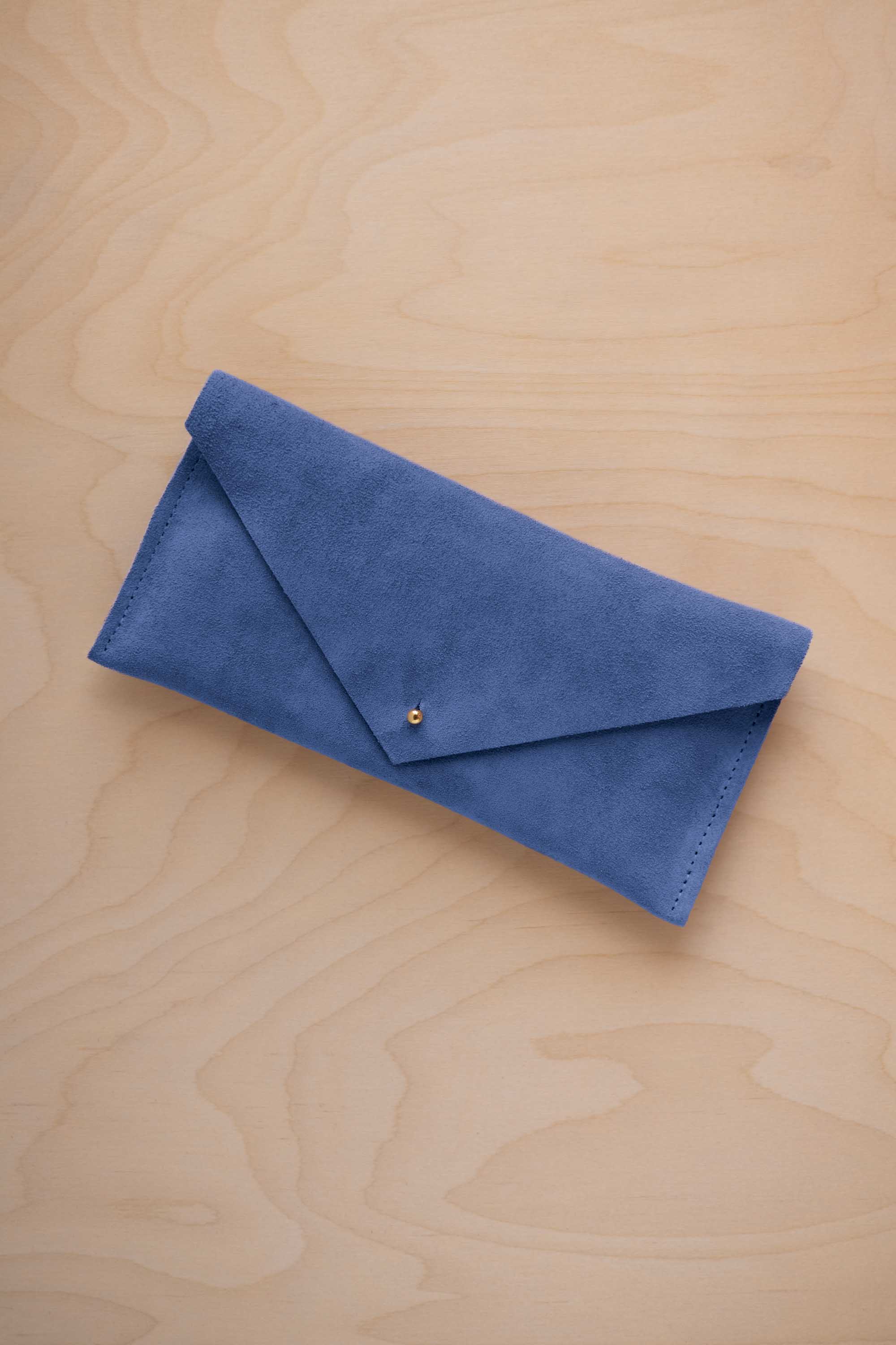 A cobalt blue suede envelope clutch wallet or bag with a small gold button clasp, resting on a light beige wood grain background.