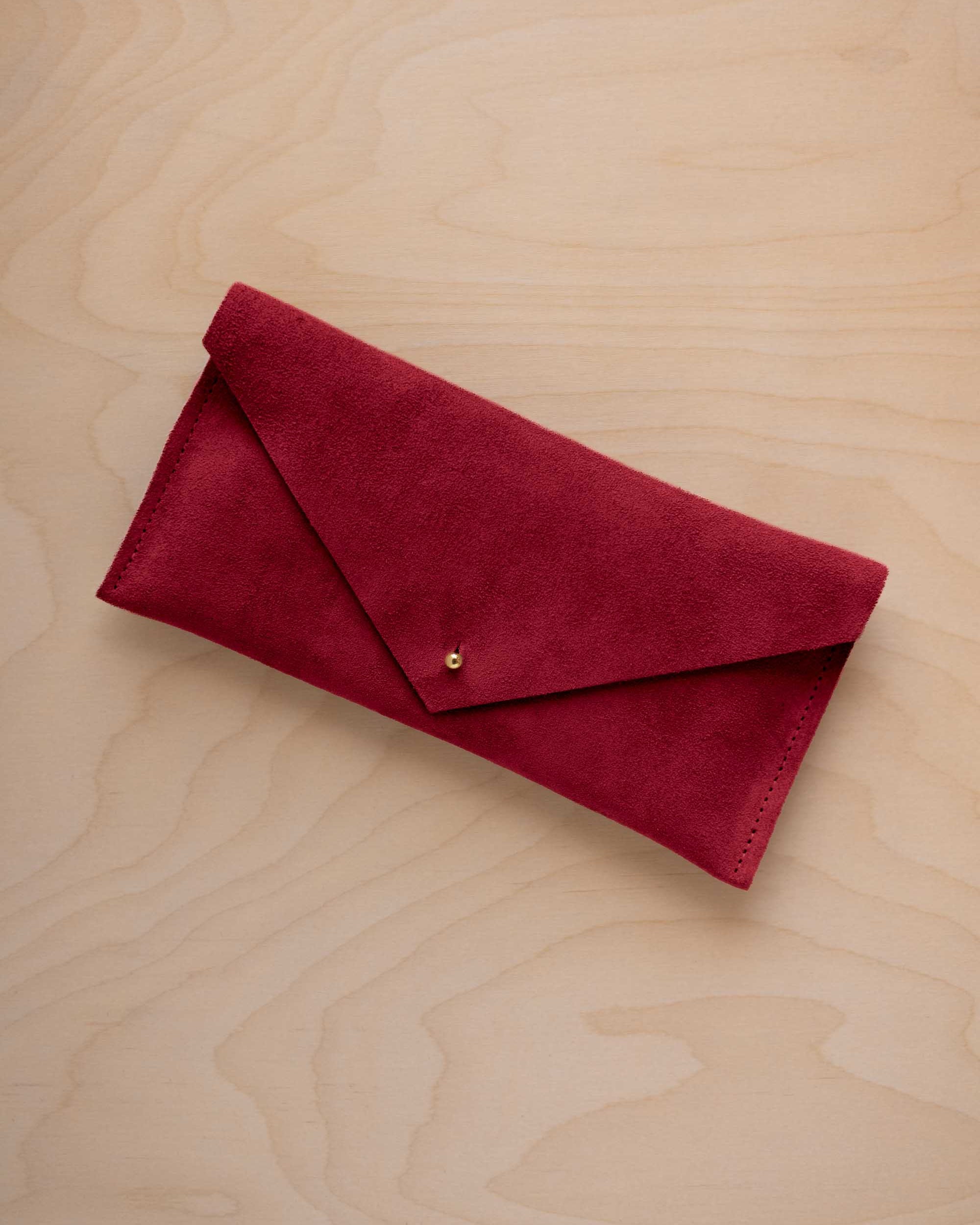 A crimson red suede envelope clutch wallet or bag with a small gold button clasp, resting on a light beige wood grain background.