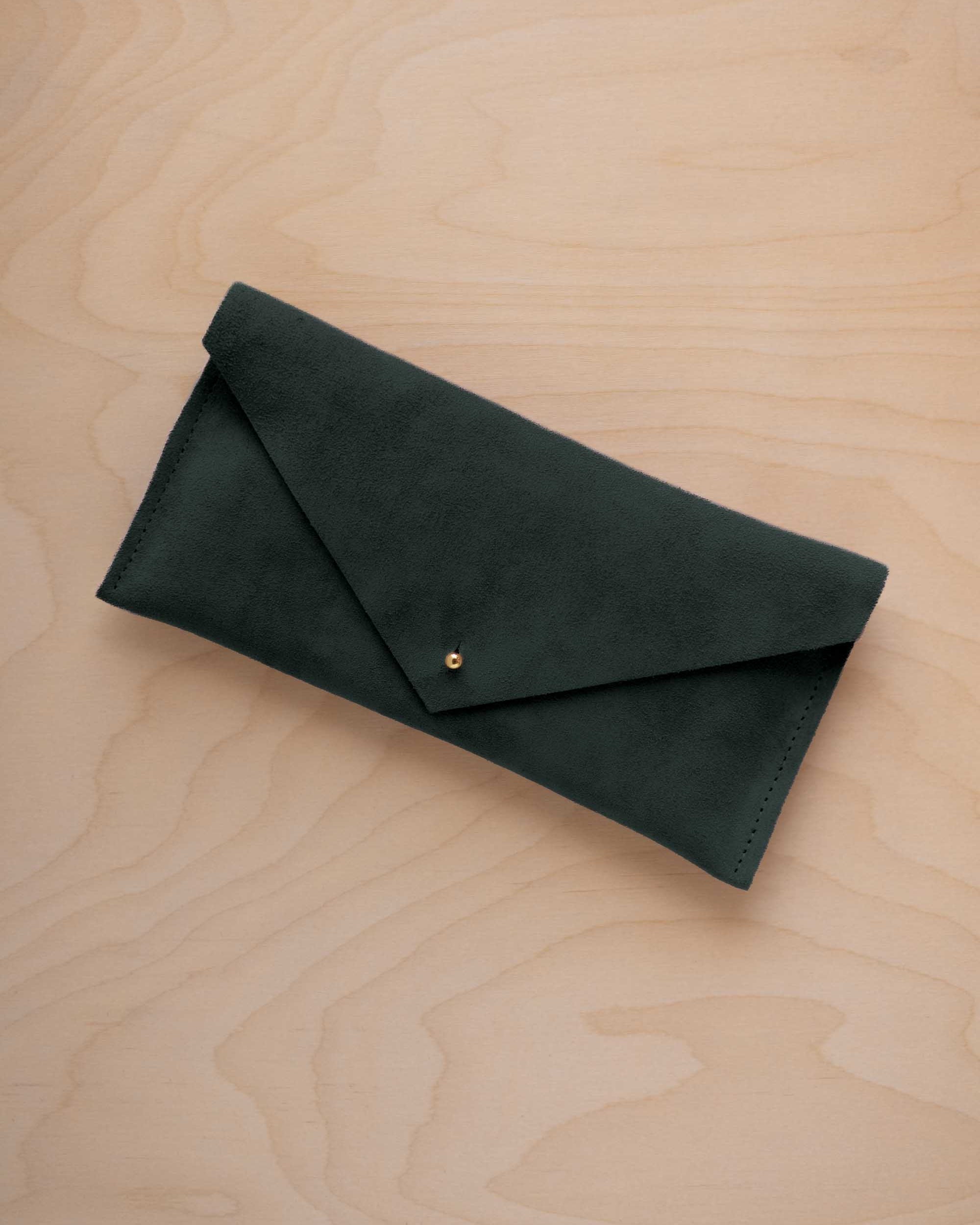 A deep forest green coloured suede envelope clutch wallet or bag with a small gold button clasp, resting on a light beige wood grain background.