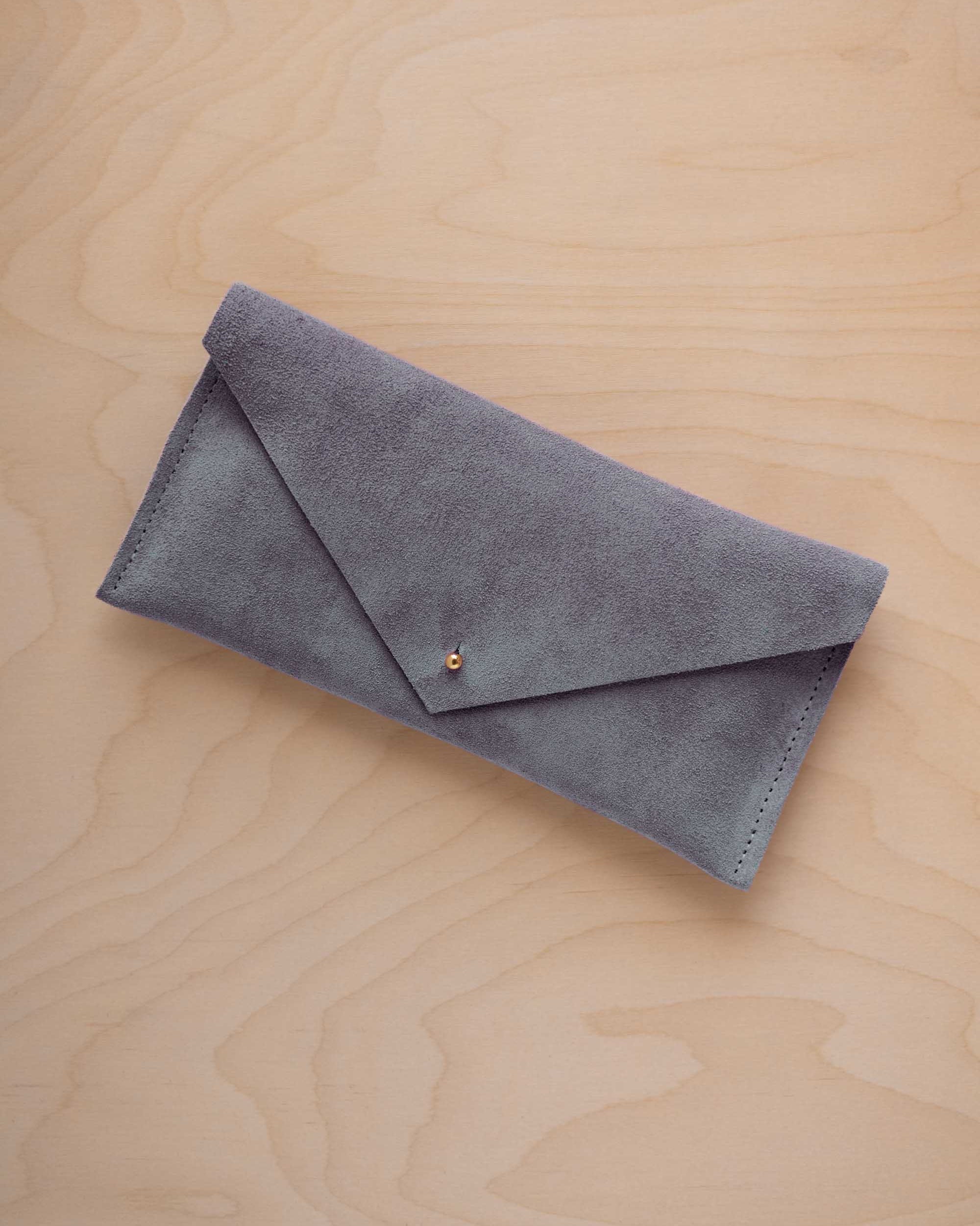 A dusky blue coloured suede envelope clutch wallet or bag with a small gold button clasp, resting on a light beige wood grain background.
