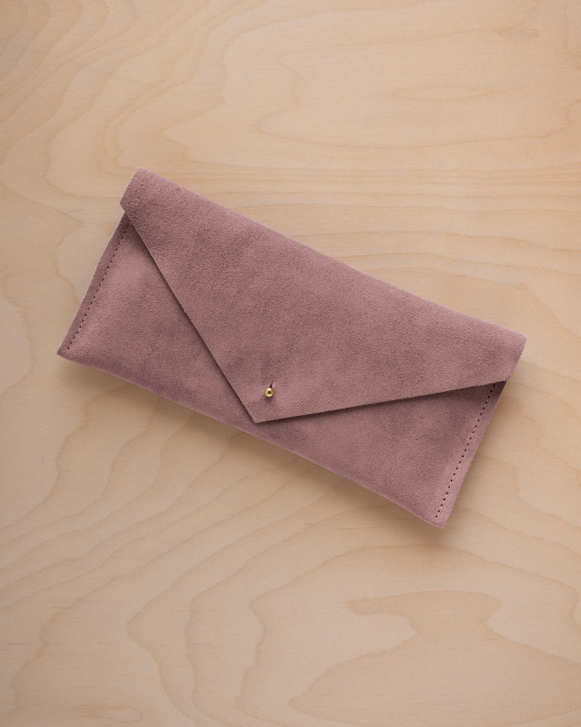A dusty mauve grey coloured suede envelope clutch wallet or bag with a small gold button clasp, resting on a light beige wood grain background.
