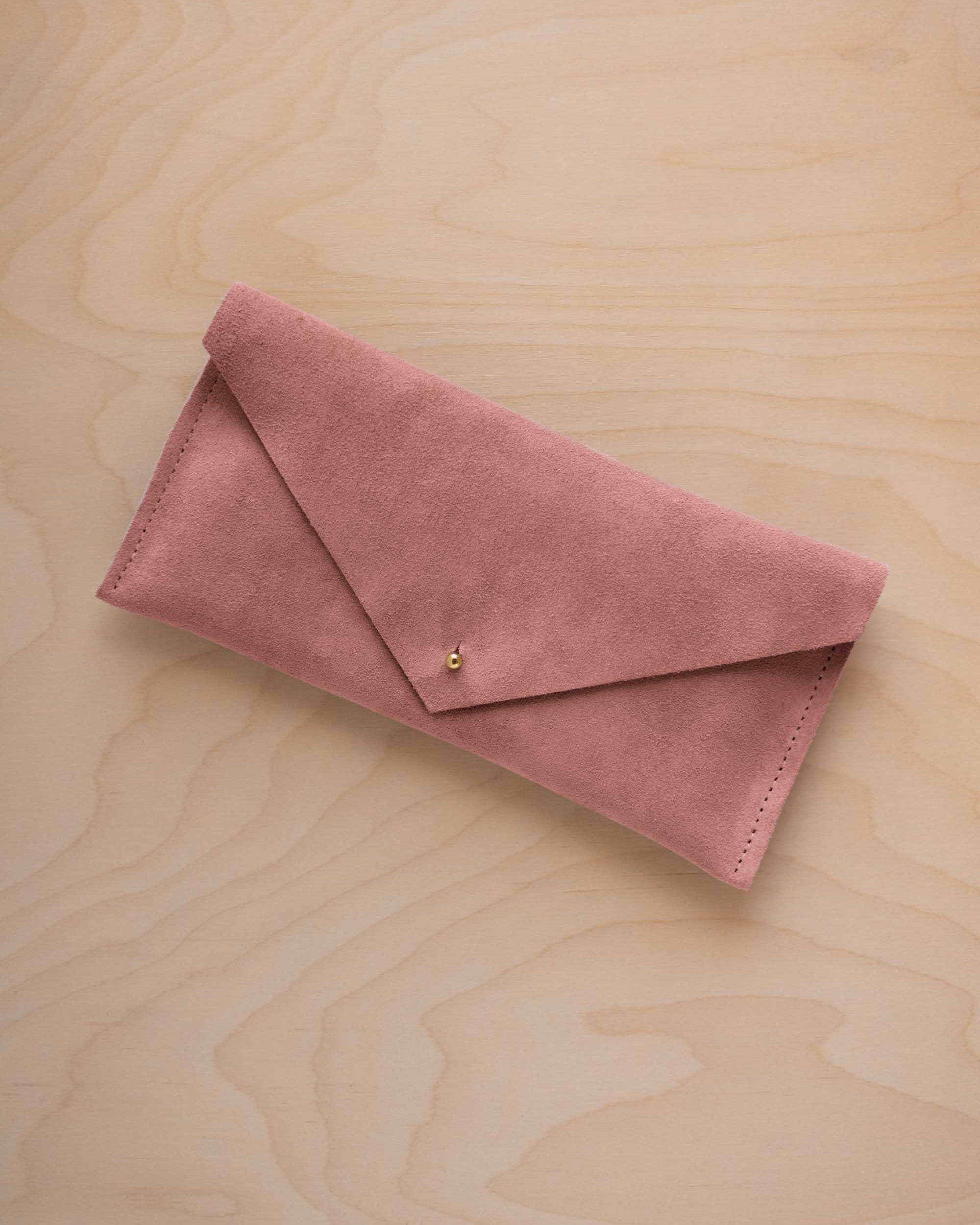 A dusty rose pink suede envelope clutch wallet or bag with a small gold button clasp, resting on a light beige wood grain background.
