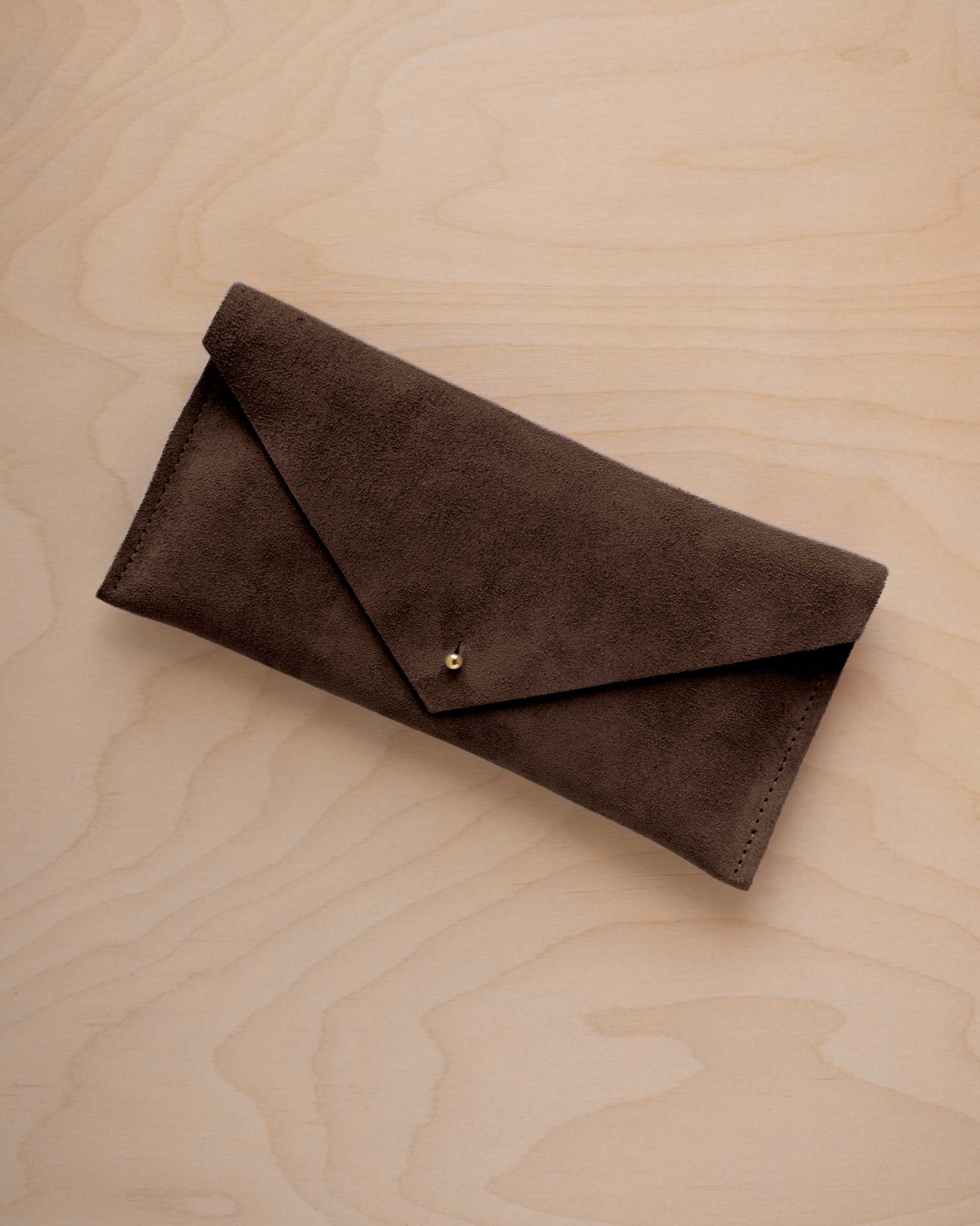 An espresso brown suede envelope clutch wallet or bag with a small gold button clasp, resting on a light beige wood grain background.