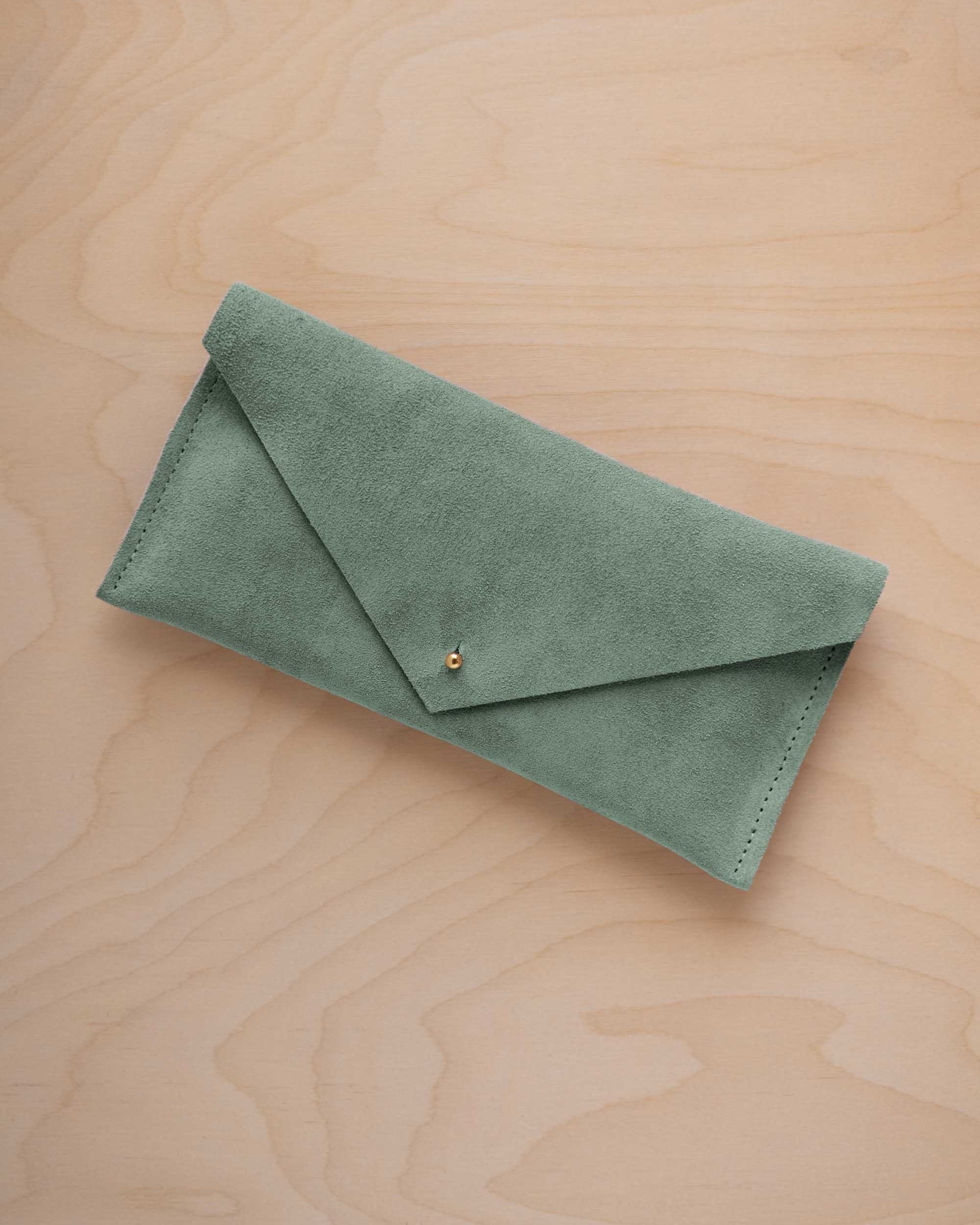 A eucalyptus sage green suede envelope clutch wallet or bag with a small gold button clasp, resting on a light beige wood grain background.