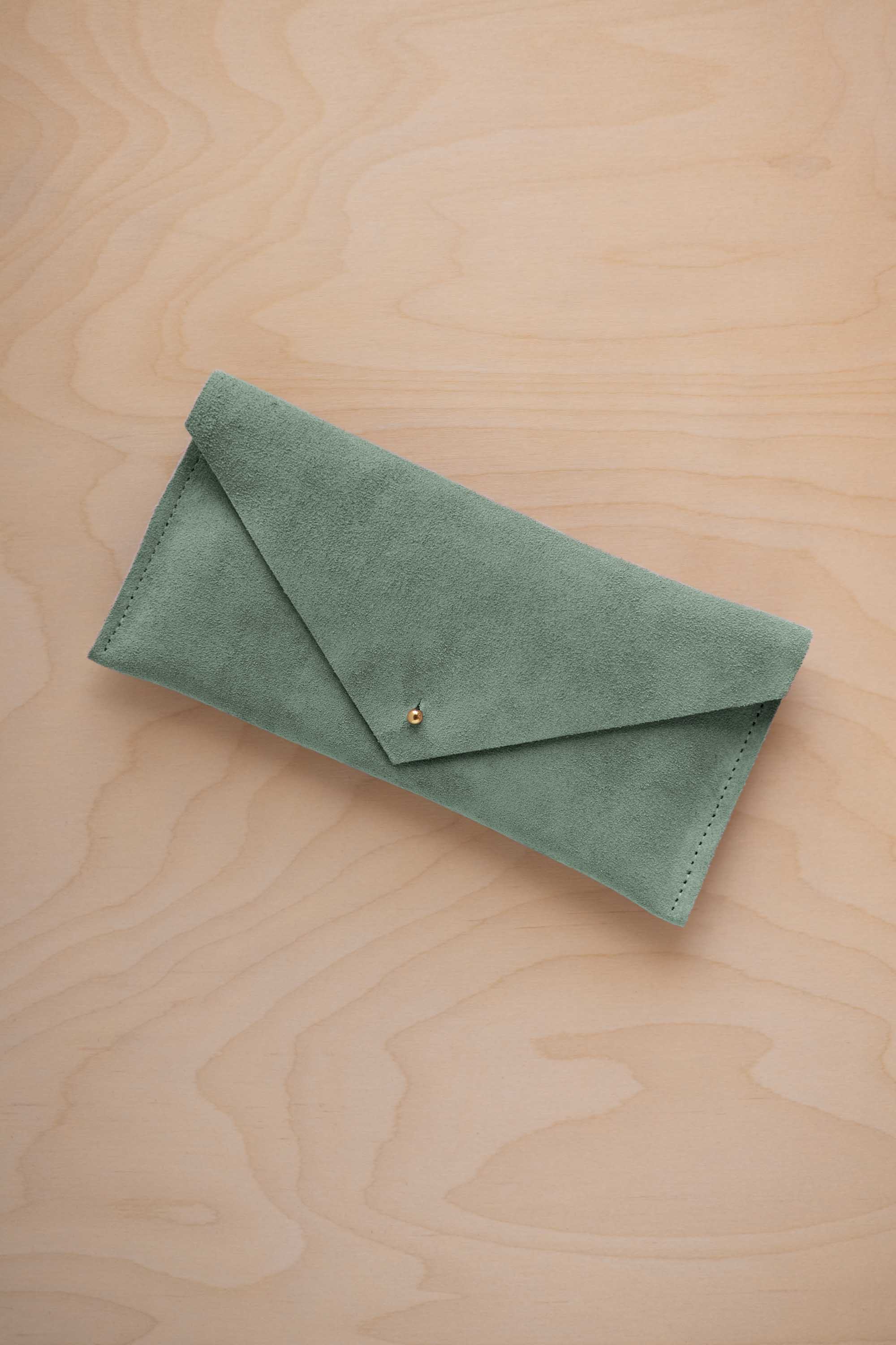A eucalyptus sage green suede envelope clutch wallet or bag with a small gold button clasp, resting on a light beige wood grain background.