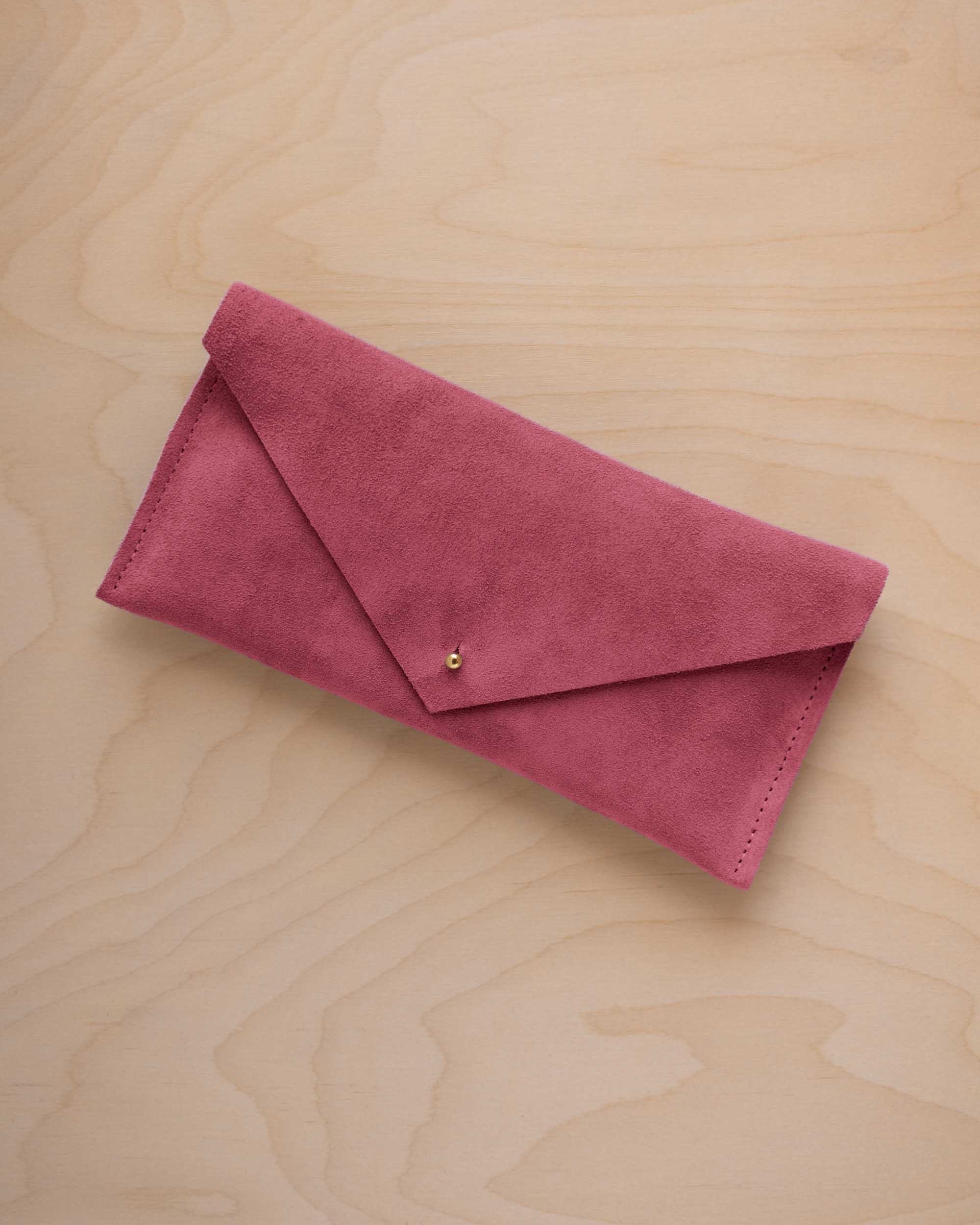 A faded rose pink suede envelope clutch wallet or bag with a small gold button clasp, resting on a light beige wood grain background.