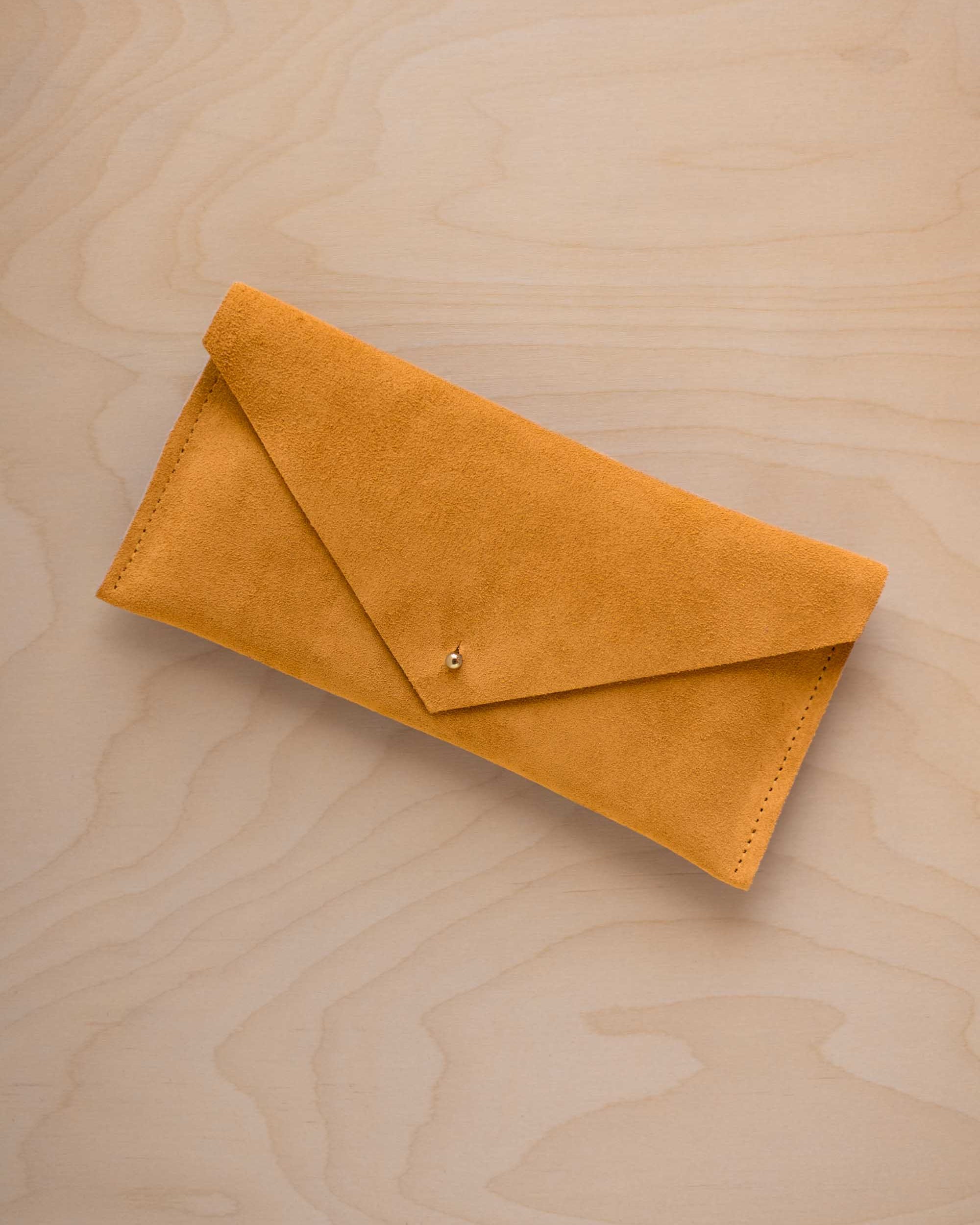 A golden ochre mustard yellow coloured envelope clutch wallet or bag with a small gold button clasp, resting on a light beige wood grain background.