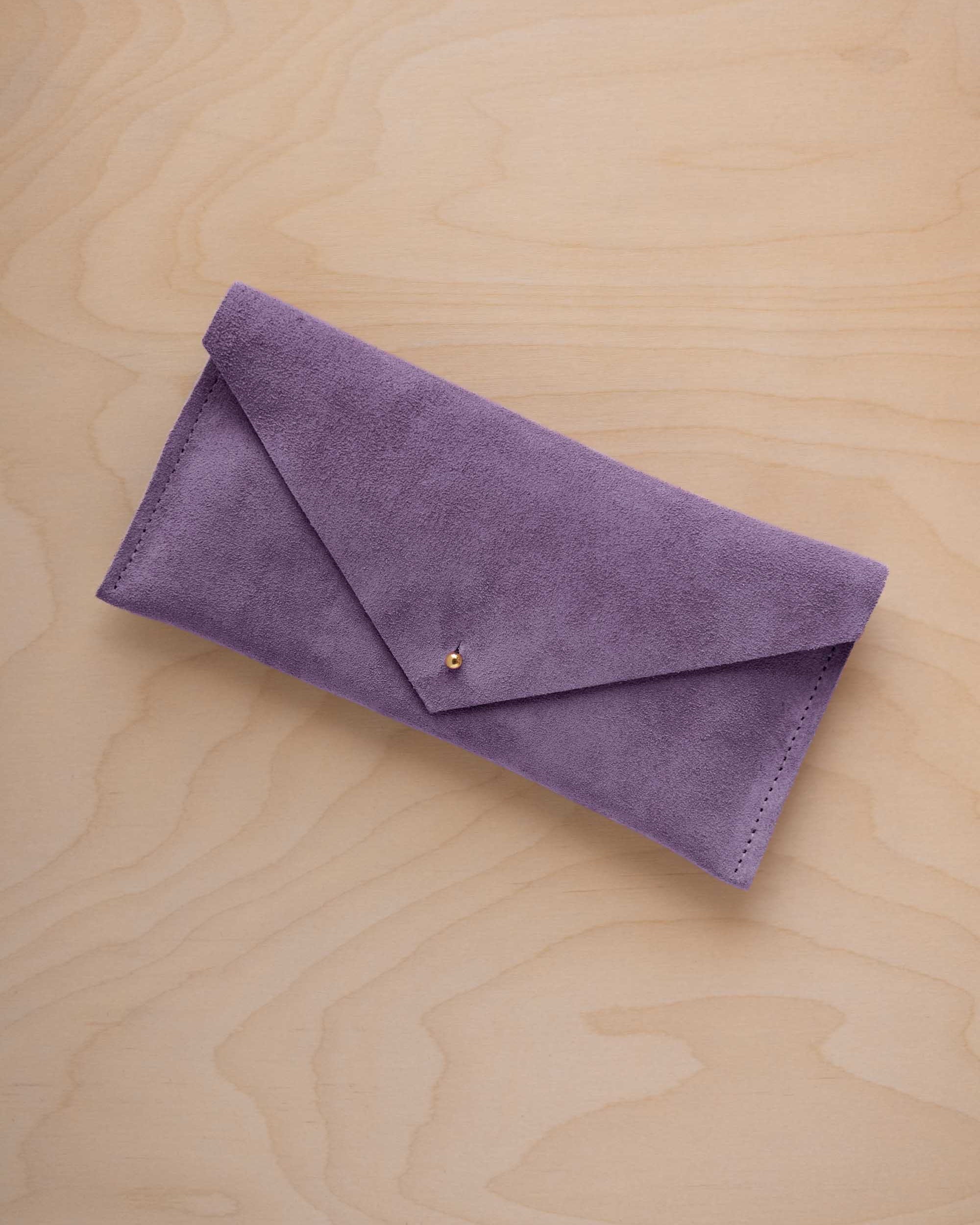 A lavender suede envelope clutch wallet or bag with a small gold button clasp, resting on a light beige wood grain background.
