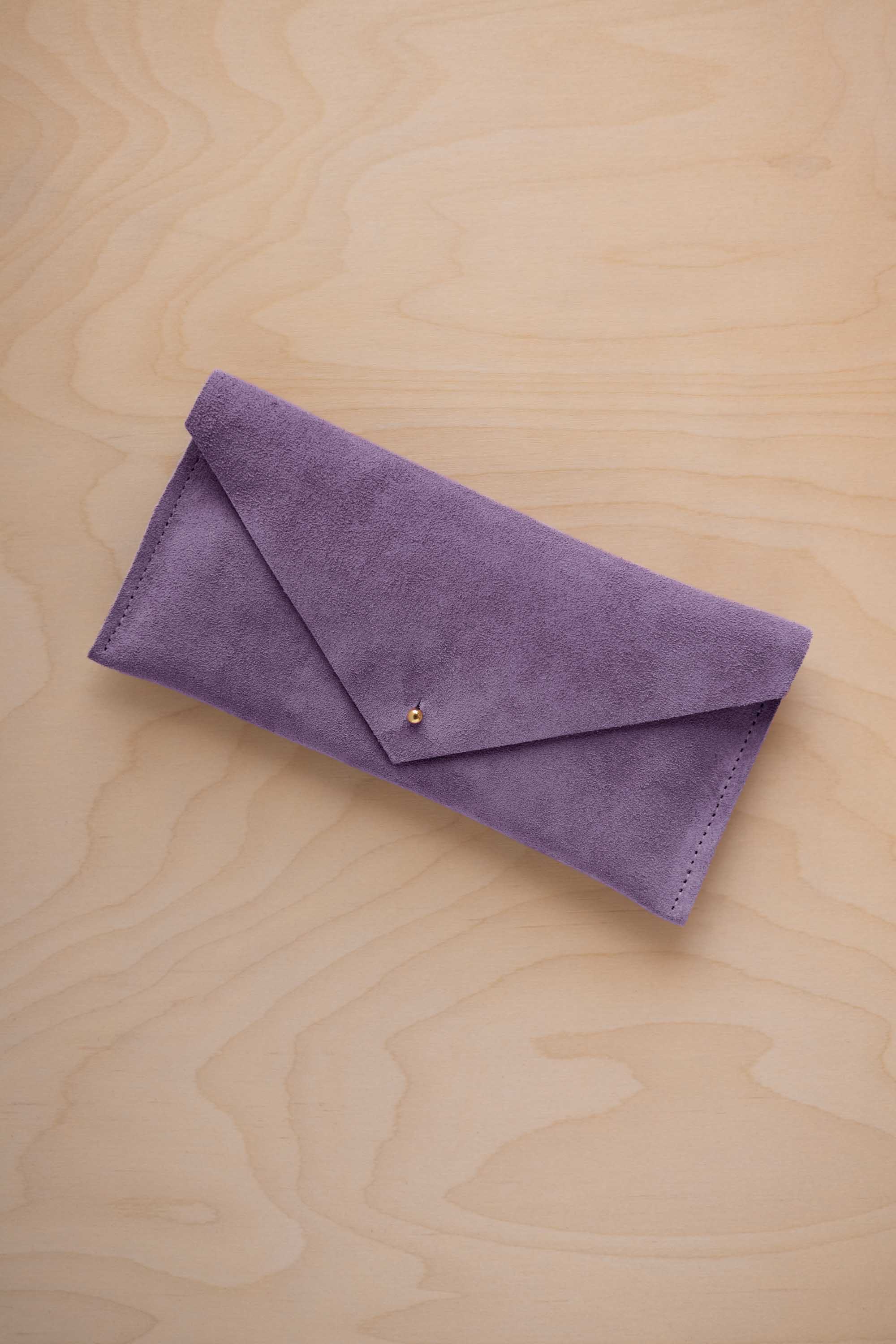 A lavender suede envelope clutch wallet or bag with a small gold button clasp, resting on a light beige wood grain background.