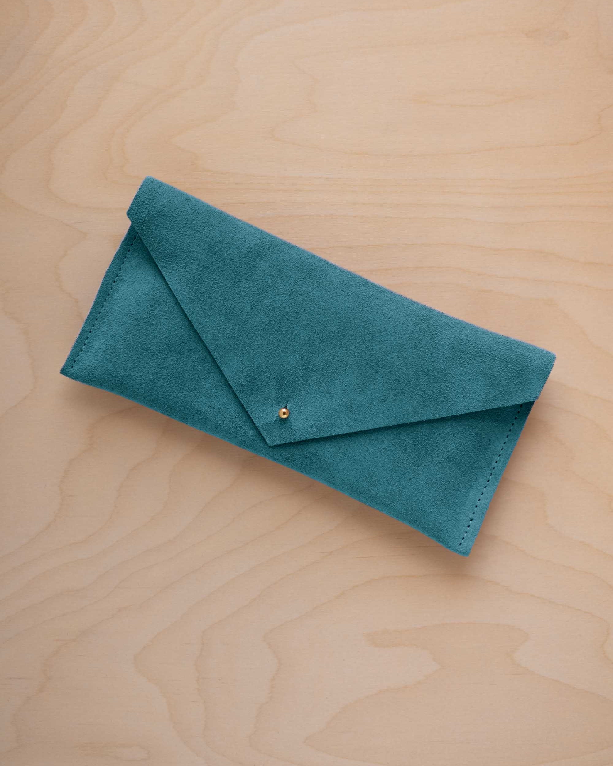 A marine teal coloured suede envelope clutch wallet or bag with a small gold button clasp, resting on a light beige wood grain background.