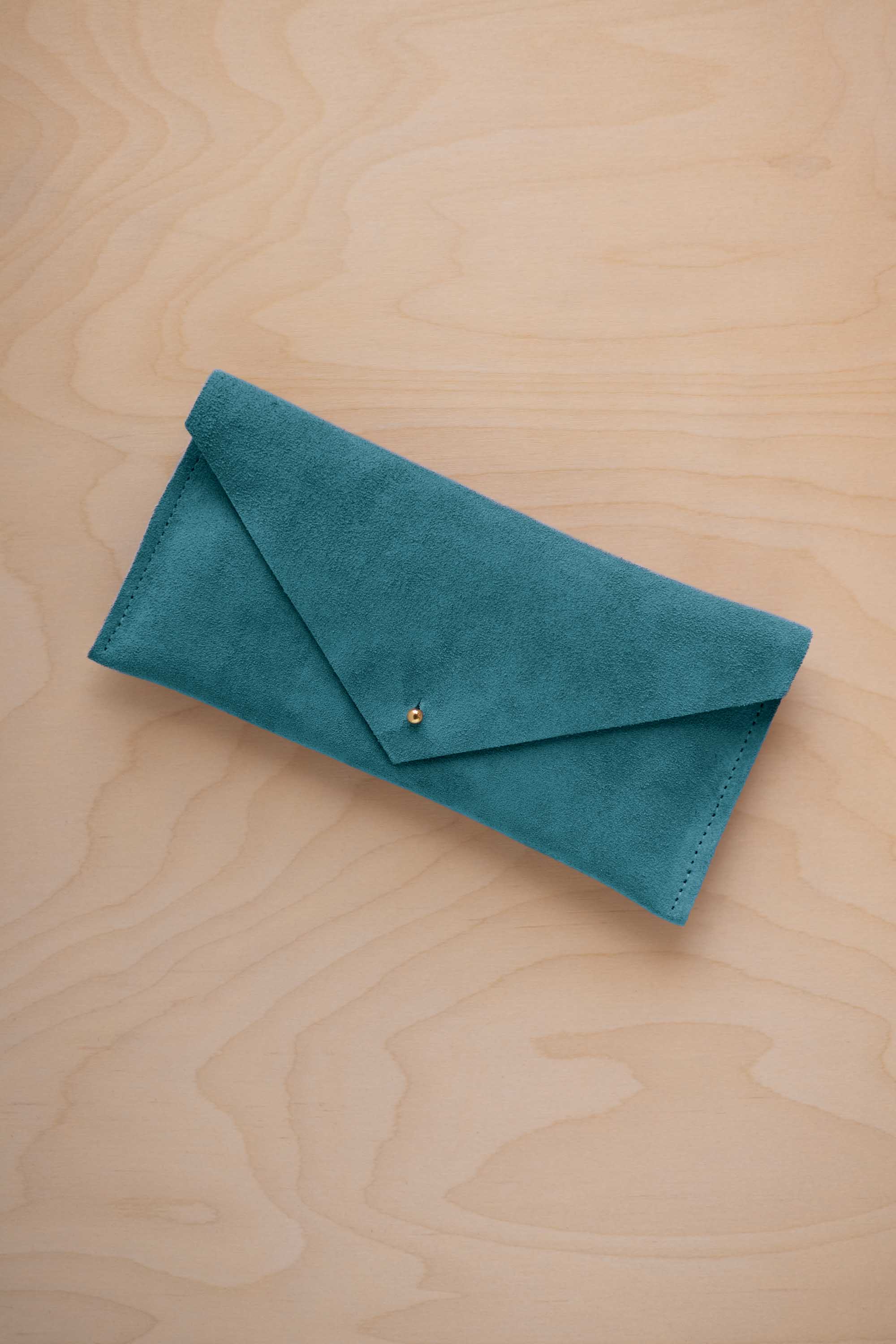 A marine teal coloured suede envelope clutch wallet or bag with a small gold button clasp, resting on a light beige wood grain background.