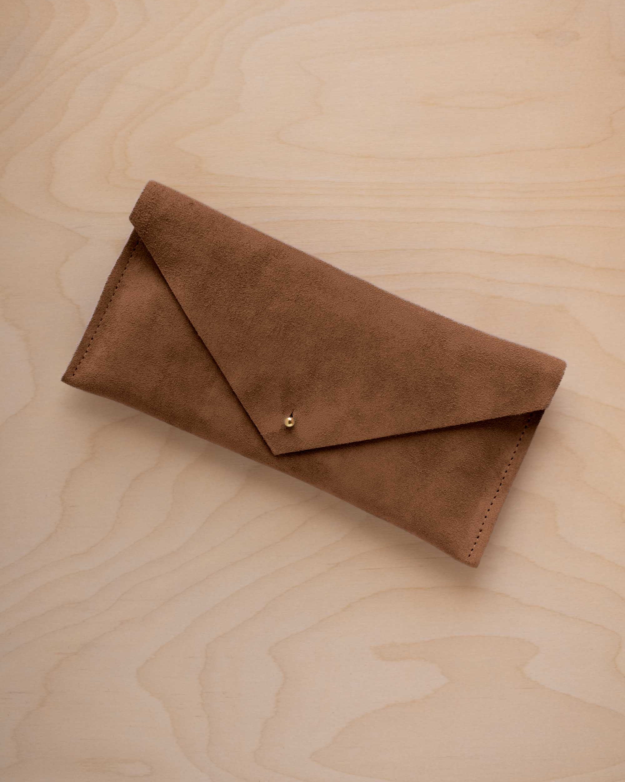 A mocha brown envelope clutch wallet or bag with a small gold button clasp, resting on a light beige wood grain background.