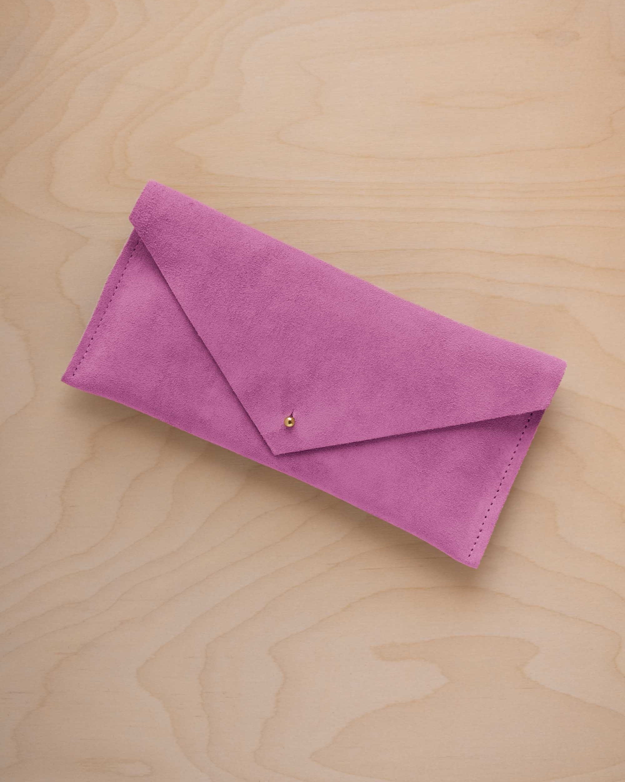 A peony pink coloured suede envelope clutch wallet or bag with a small gold button clasp, resting on a light beige wood grain background.
