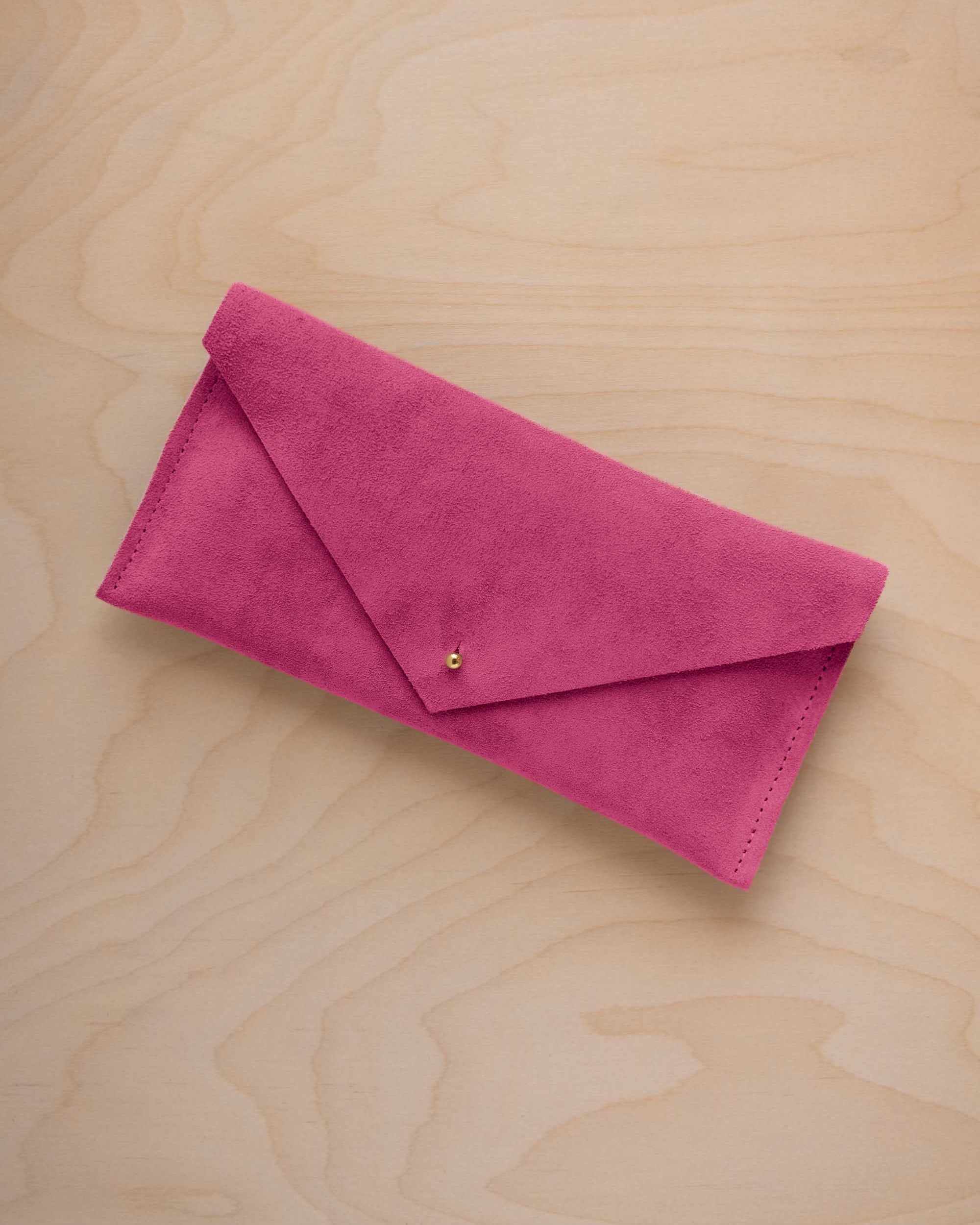 A raspberry pink coloured suede envelope clutch wallet or bag with a small gold button clasp, resting on a light beige wood grain background.