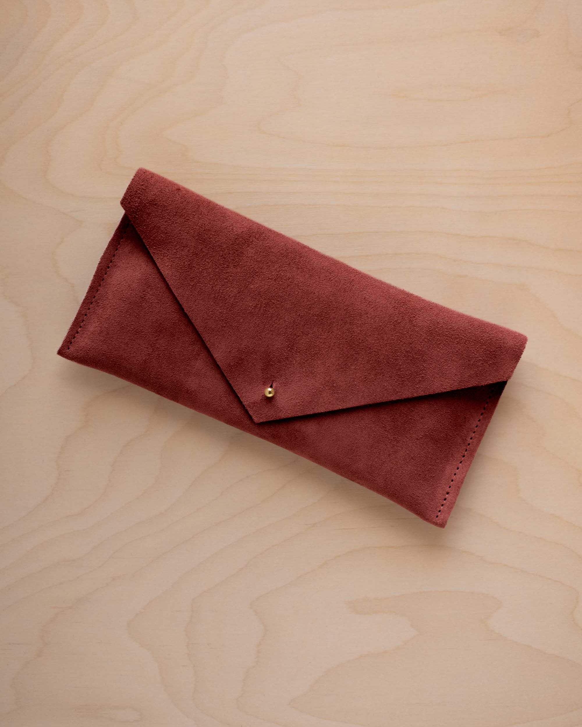 A rust red coloured suede envelope clutch wallet or bag with a small gold button clasp, resting on a light beige wood grain background.