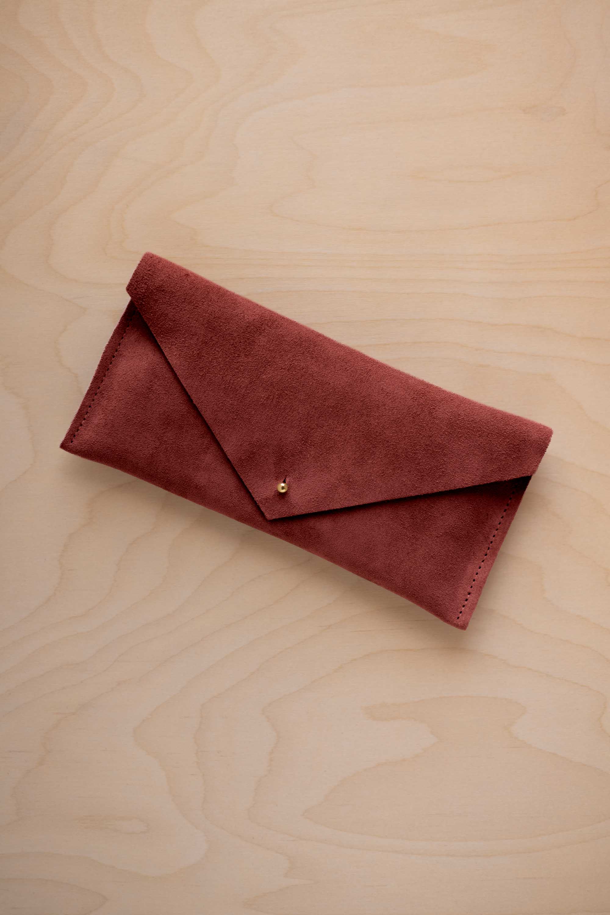 A rust red coloured suede envelope clutch wallet or bag with a small gold button clasp, resting on a light beige wood grain background.