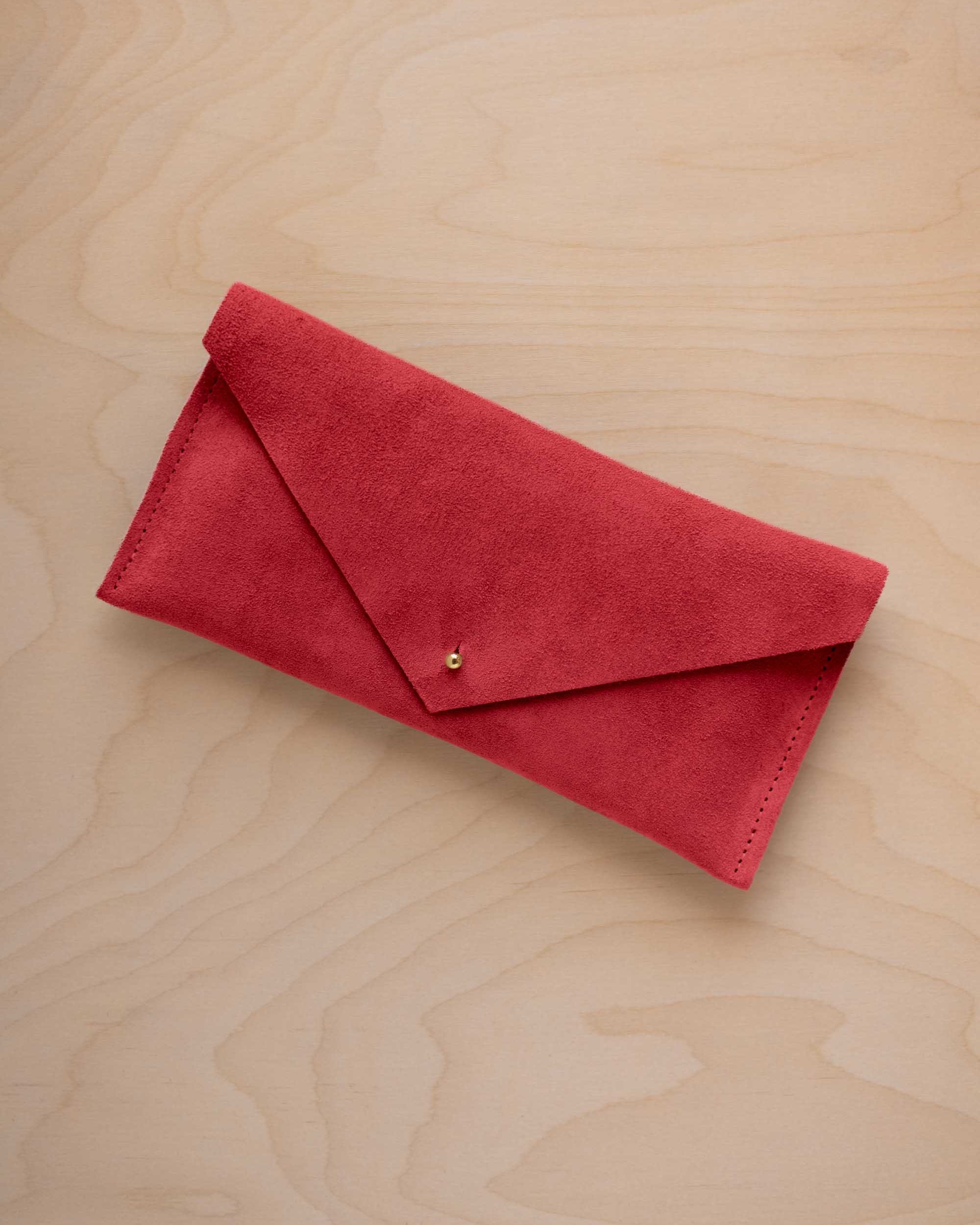 A scarlet red suede envelope clutch wallet or bag with a small gold button clasp, resting on a light beige wood grain background.