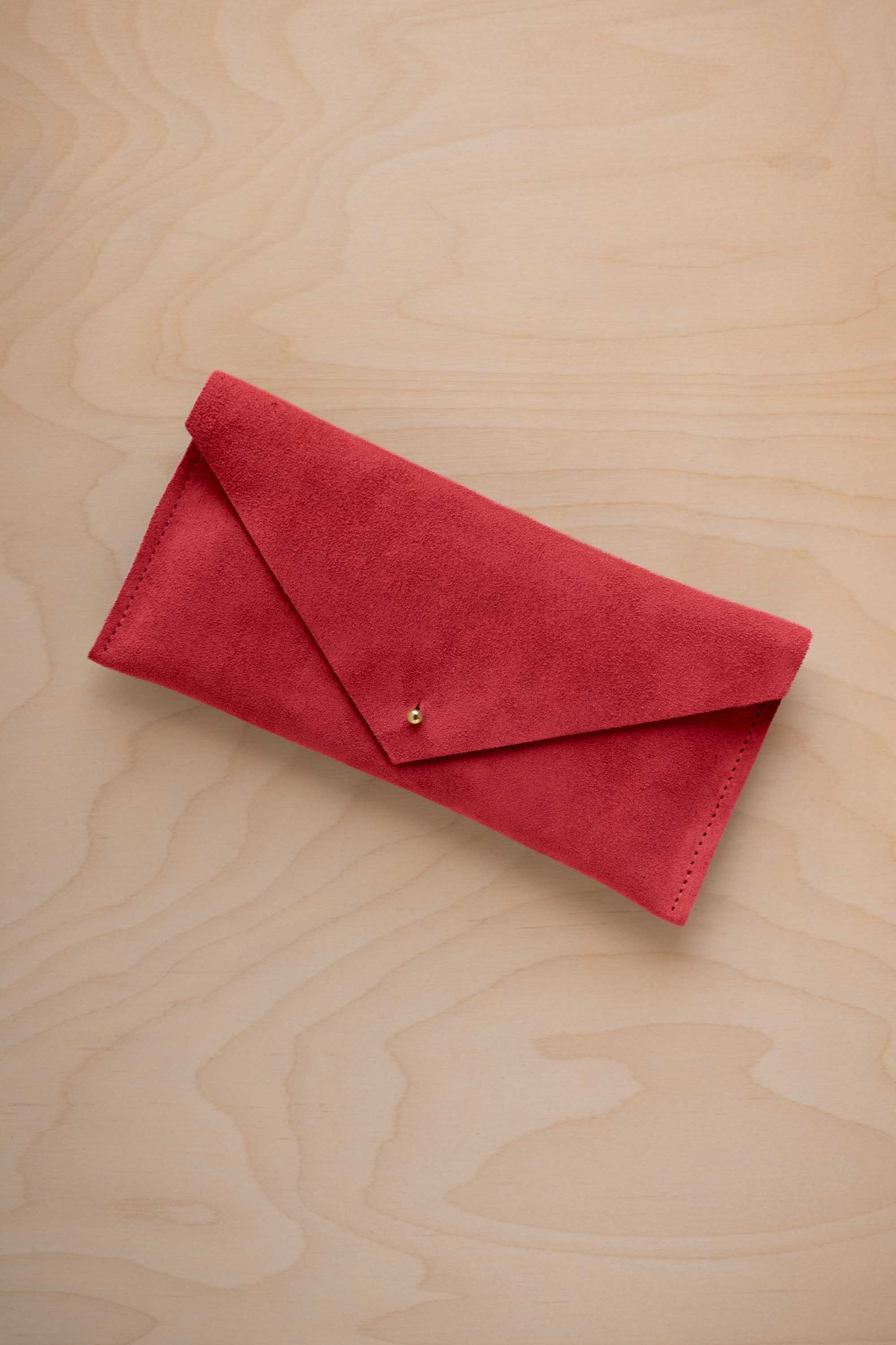 A scarlet red suede envelope clutch wallet or bag with a small gold button clasp, resting on a light beige wood grain background.