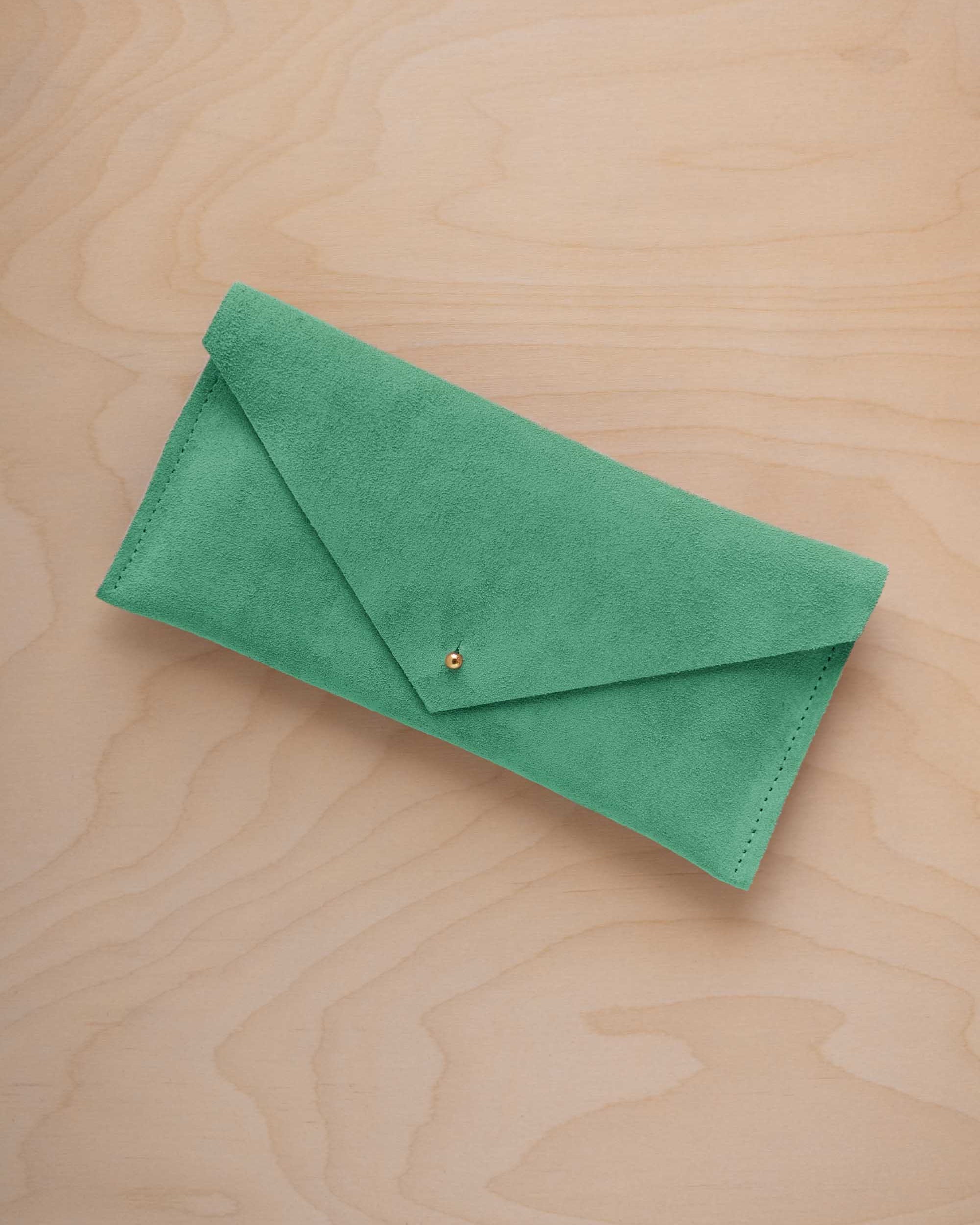 A seafoam turquoise green coloured suede envelope clutch wallet or bag with a small gold button clasp, resting on a light beige wood grain background.