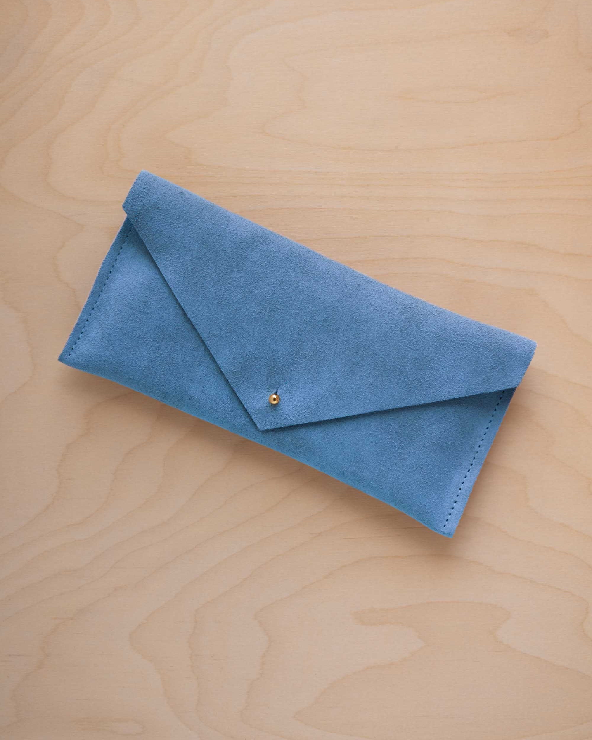 A sky blue coloured suede envelope clutch wallet or bag with a small gold button clasp, resting on a light beige wood grain background.