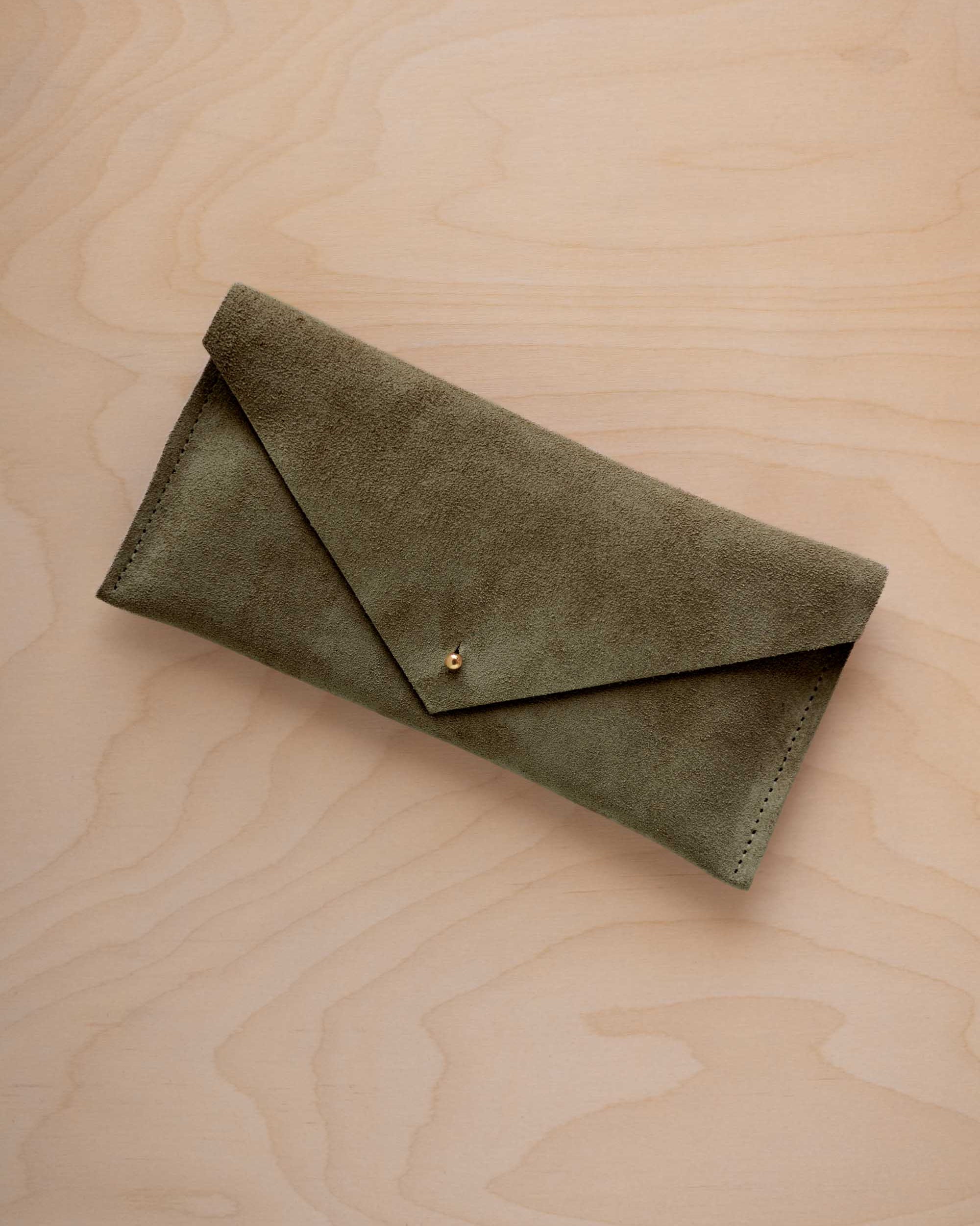 A soft olive green coloured suede envelope clutch wallet or bag with a small gold button clasp, resting on a light beige wood grain background.