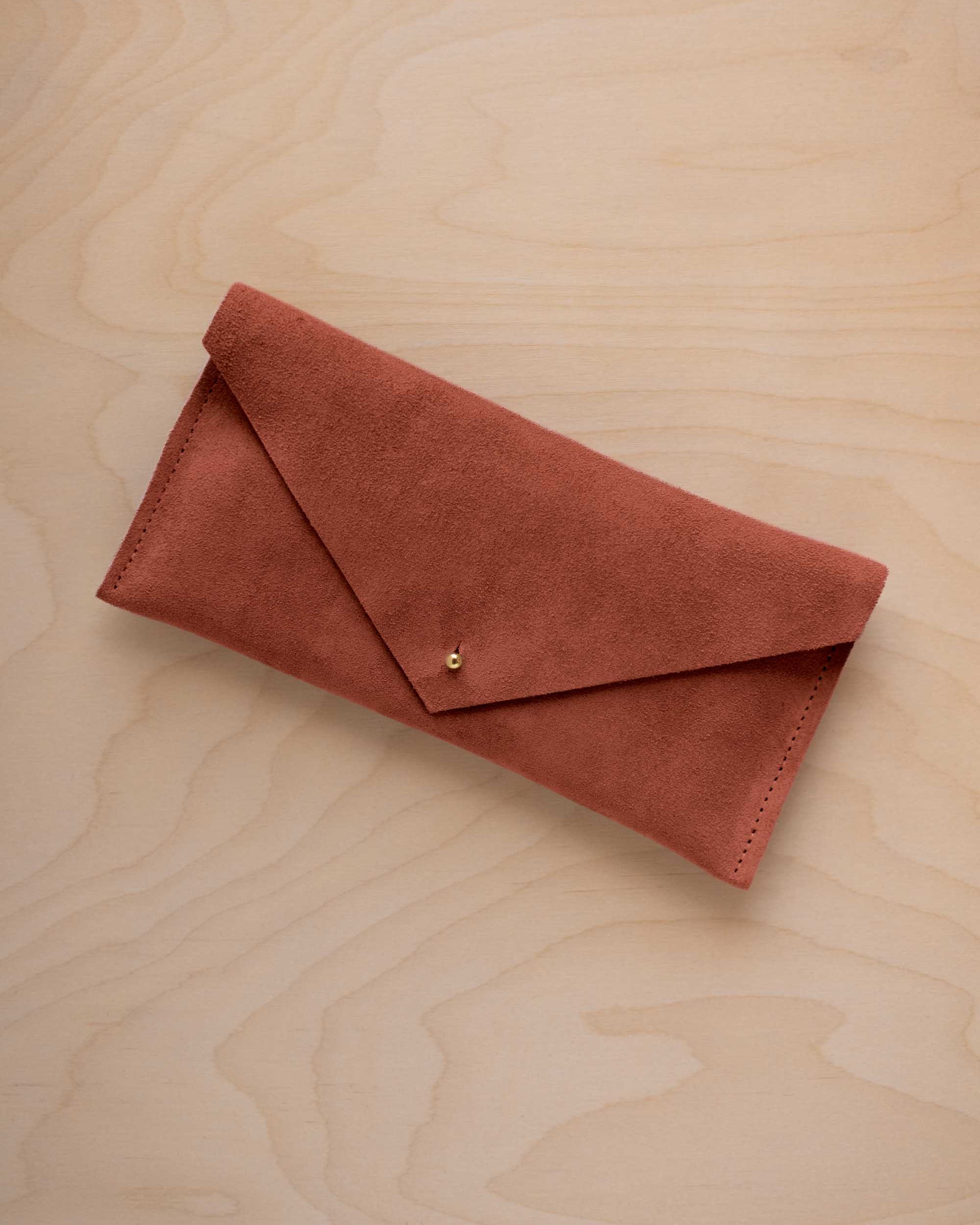 A terracotta coloured envelope clutch wallet or bag with a small gold button clasp, resting on a light beige wood grain background.