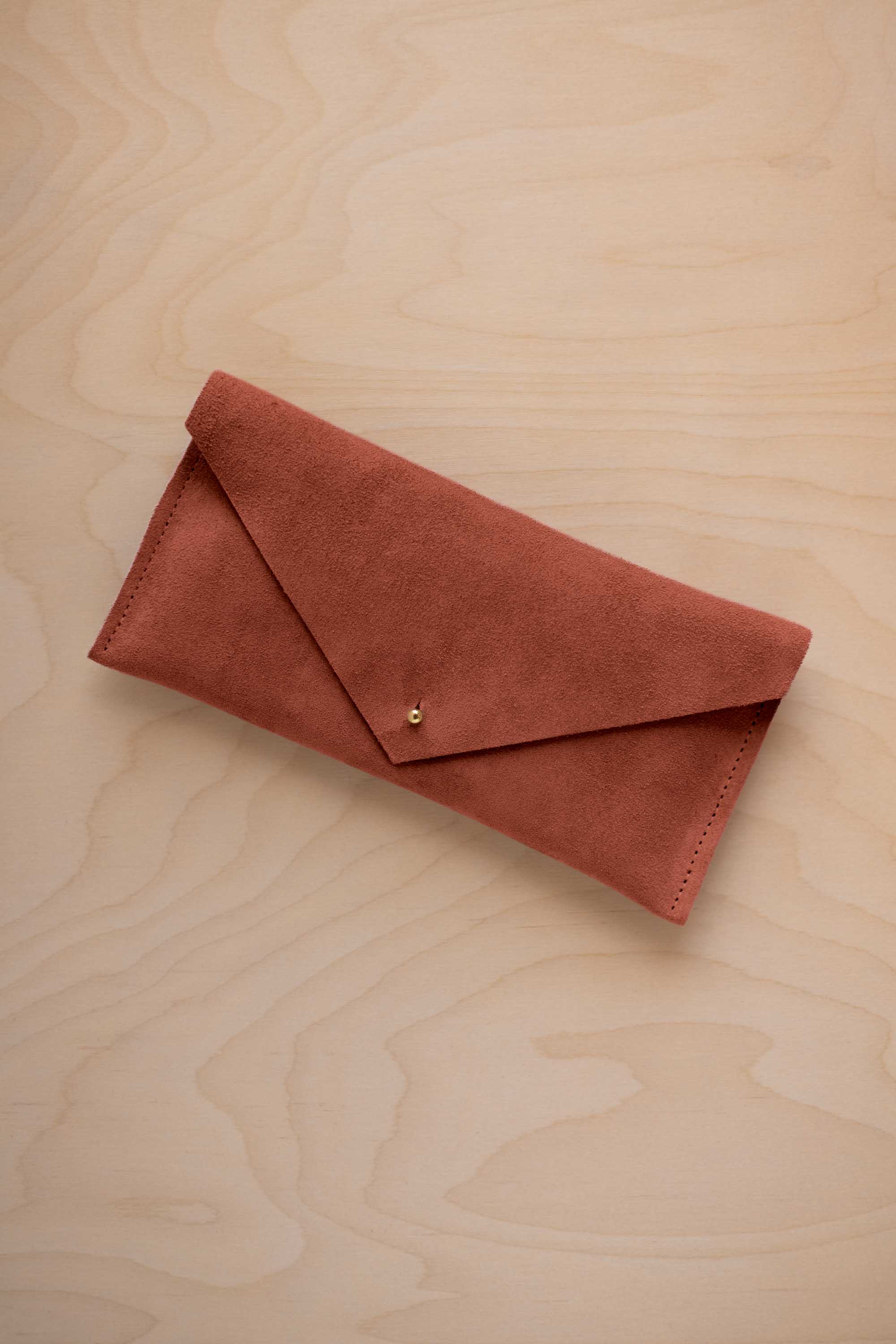 A terracotta coloured envelope clutch wallet or bag with a small gold button clasp, resting on a light beige wood grain background.