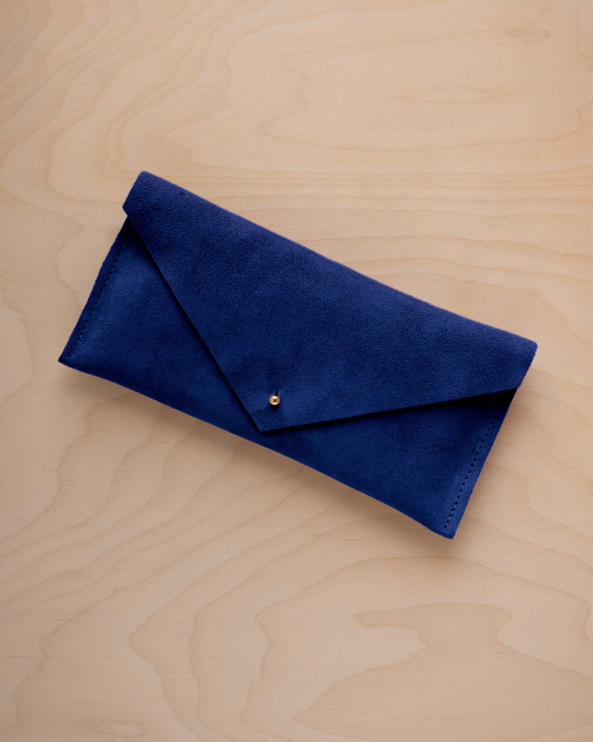 An ultramarine blue suede envelope clutch wallet or bag with a small gold button clasp, resting on a light beige wood grain background.