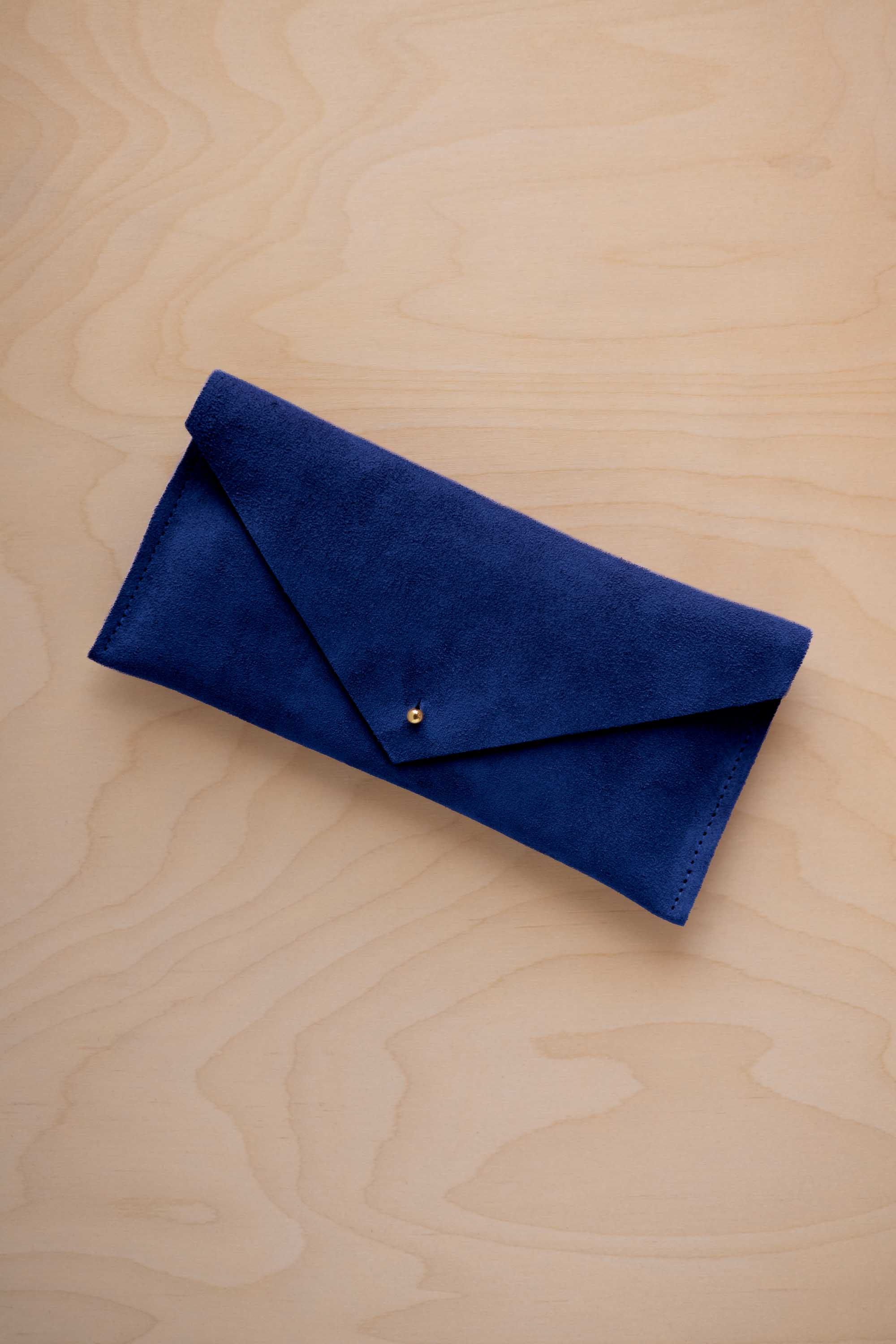 An ultramarine blue suede envelope clutch wallet or bag with a small gold button clasp, resting on a light beige wood grain background.