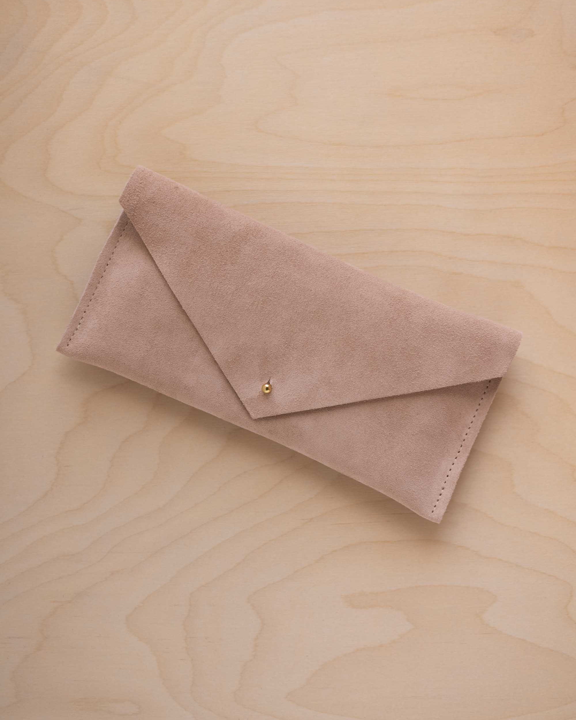 A beige suede envelope clutch wallet or bag with a small gold button clasp, resting on a light beige wood grain background.