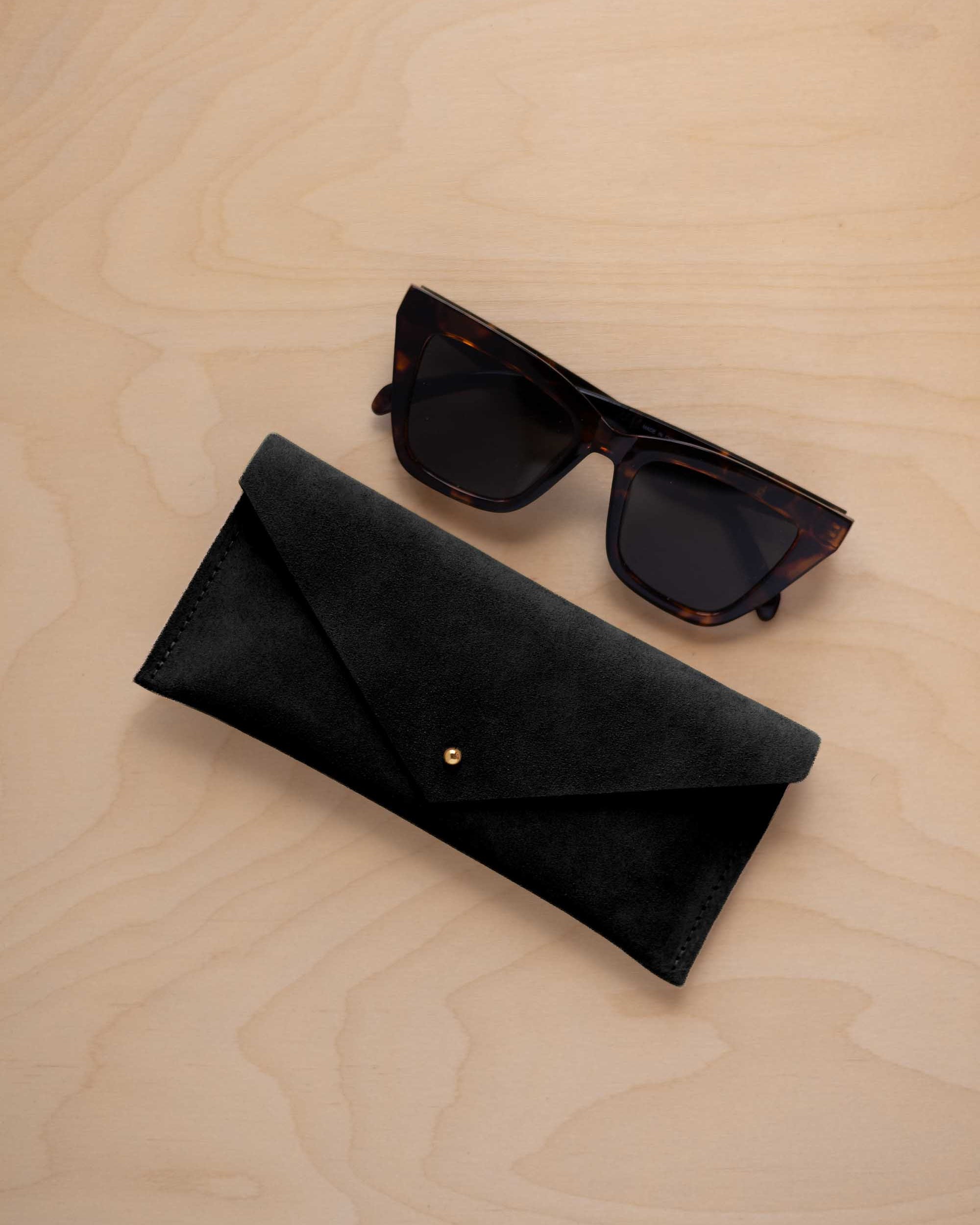 A pair of dark tortoiseshell sunglasses sit next to a black suede envelope style eye glasses case with a small gold button, set on a light wood grain background.