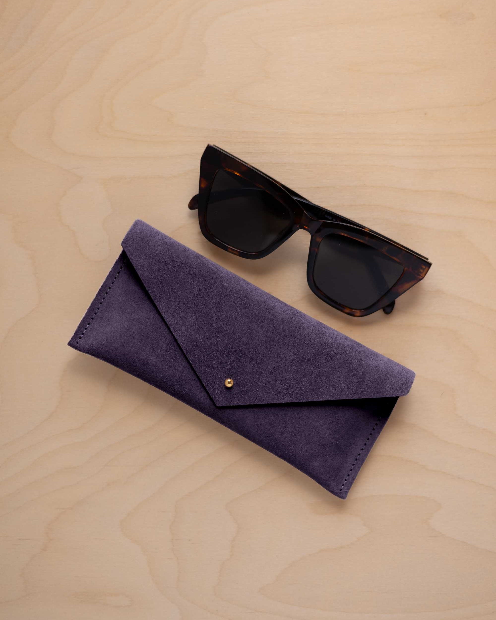 A pair of dark tortoiseshell sunglasses sit next to a blackcurrent dark purple suede envelope style eye glasses case with a small gold button, set on a light wood grain background.