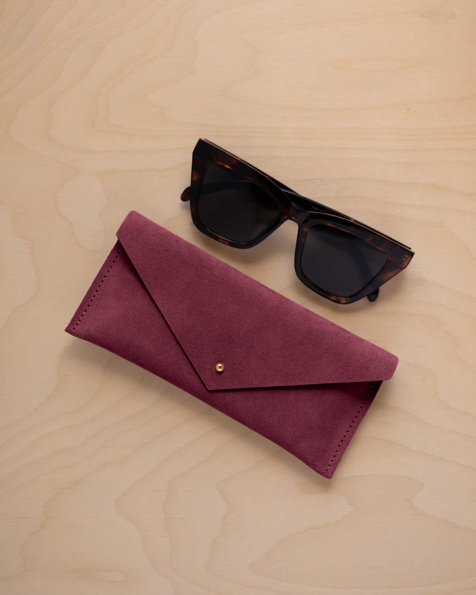 A pair of dark tortoiseshell sunglasses sit next to a burgundy suede envelope style eye glasses case with a small gold button, set on a light wood grain background.