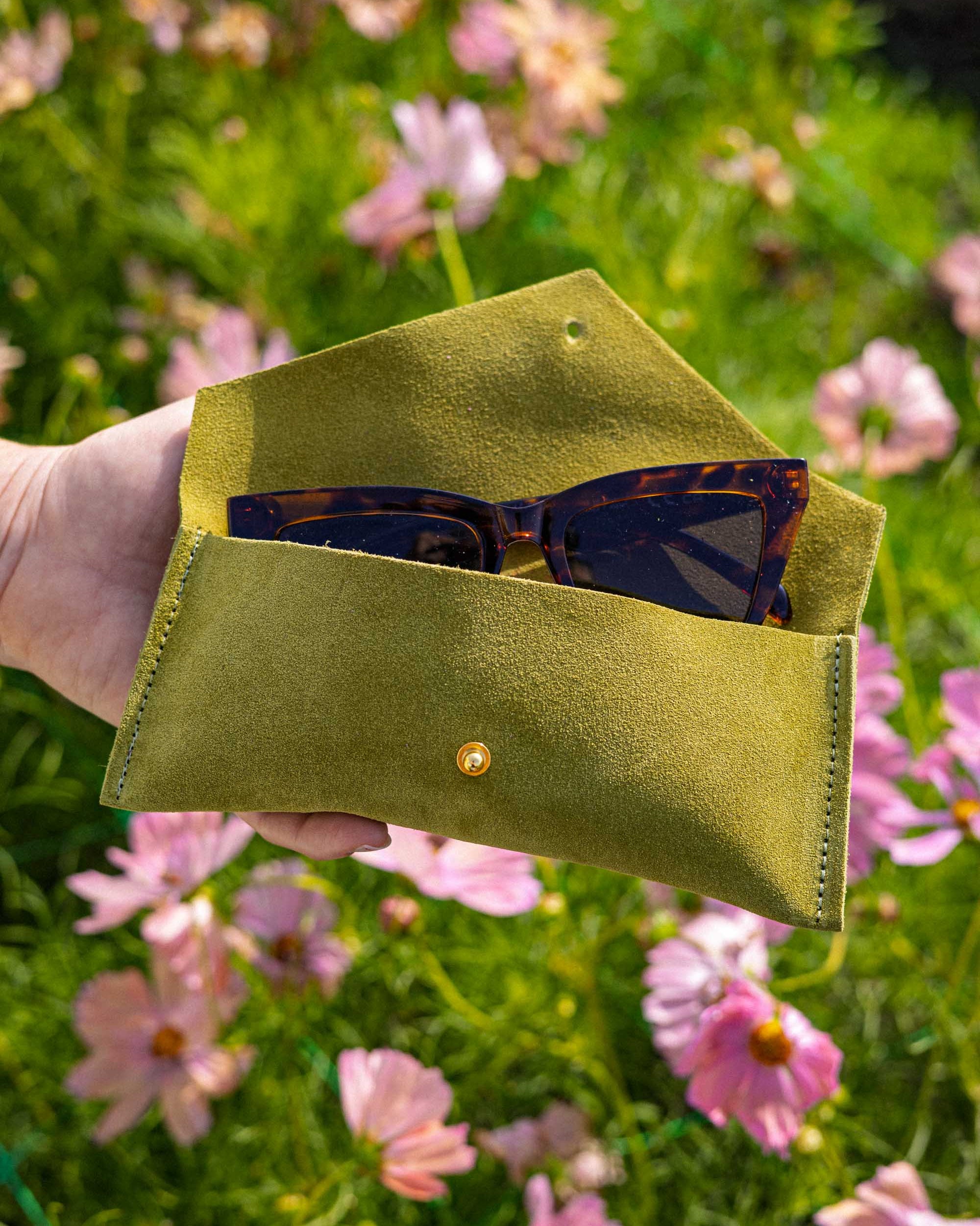 A hand holds an open chartreuse green suede sunglasses case with a small gold button, containing tortoiseshell sunglasses, set against a blurred background of pink flowers and green grass.