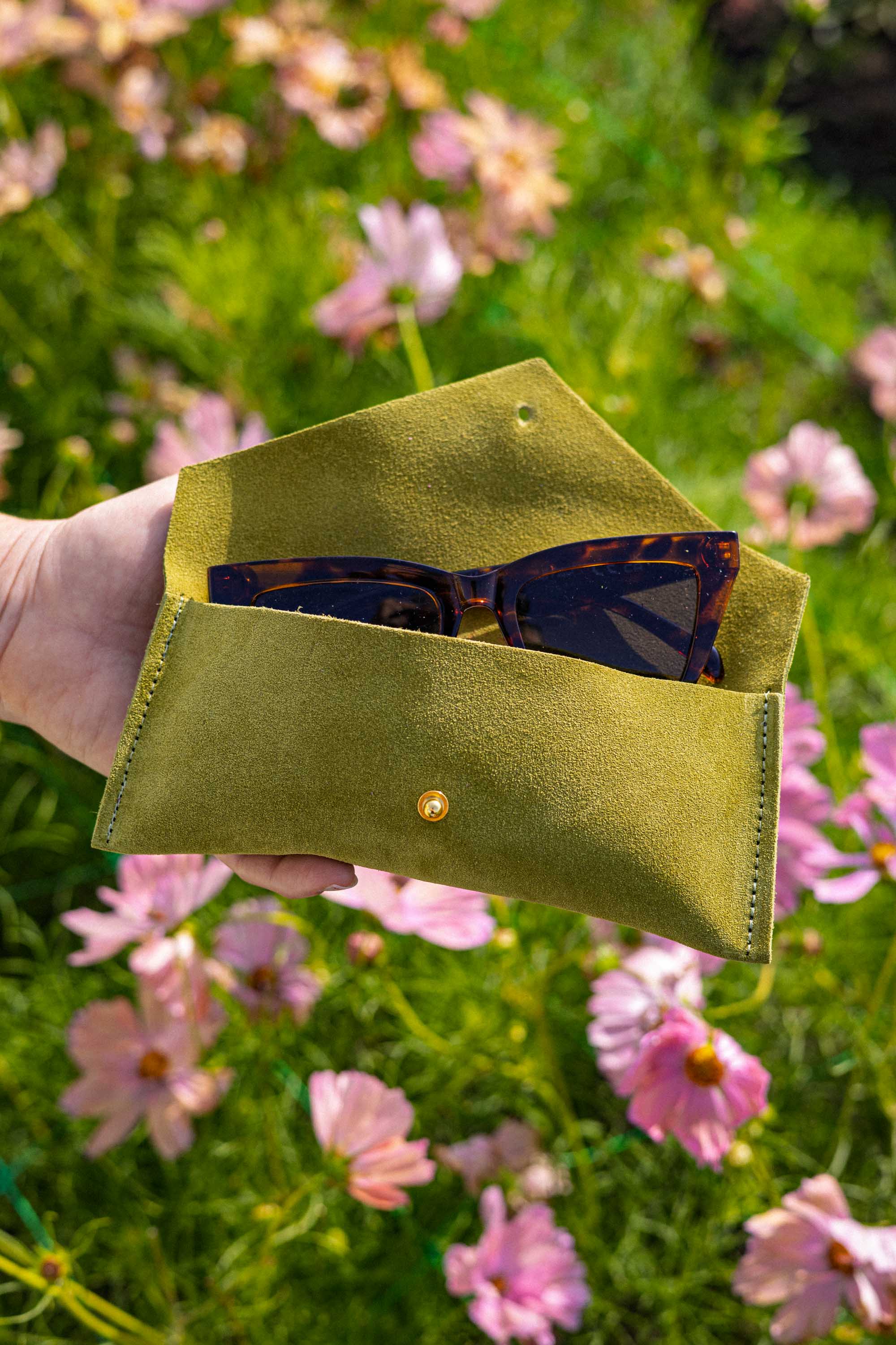 A hand holds an open chartreuse green suede sunglasses case with a small gold button, containing tortoiseshell sunglasses, set against a blurred background of pink flowers and green grass.