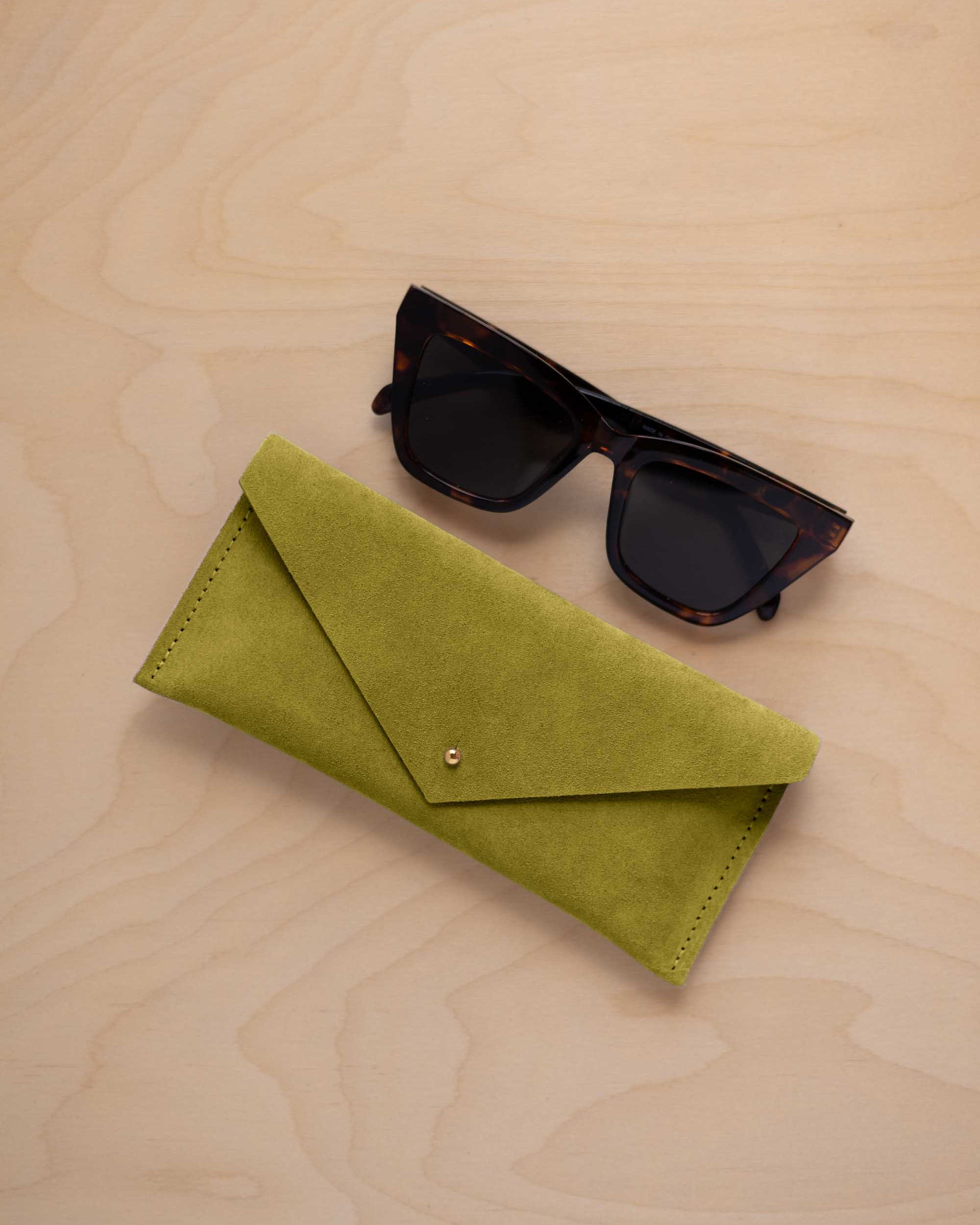 A pair of dark tortoiseshell sunglasses sit next to a chartreuse lime green suede envelope style eye glasses case with a small gold button, set on a light wood grain background.