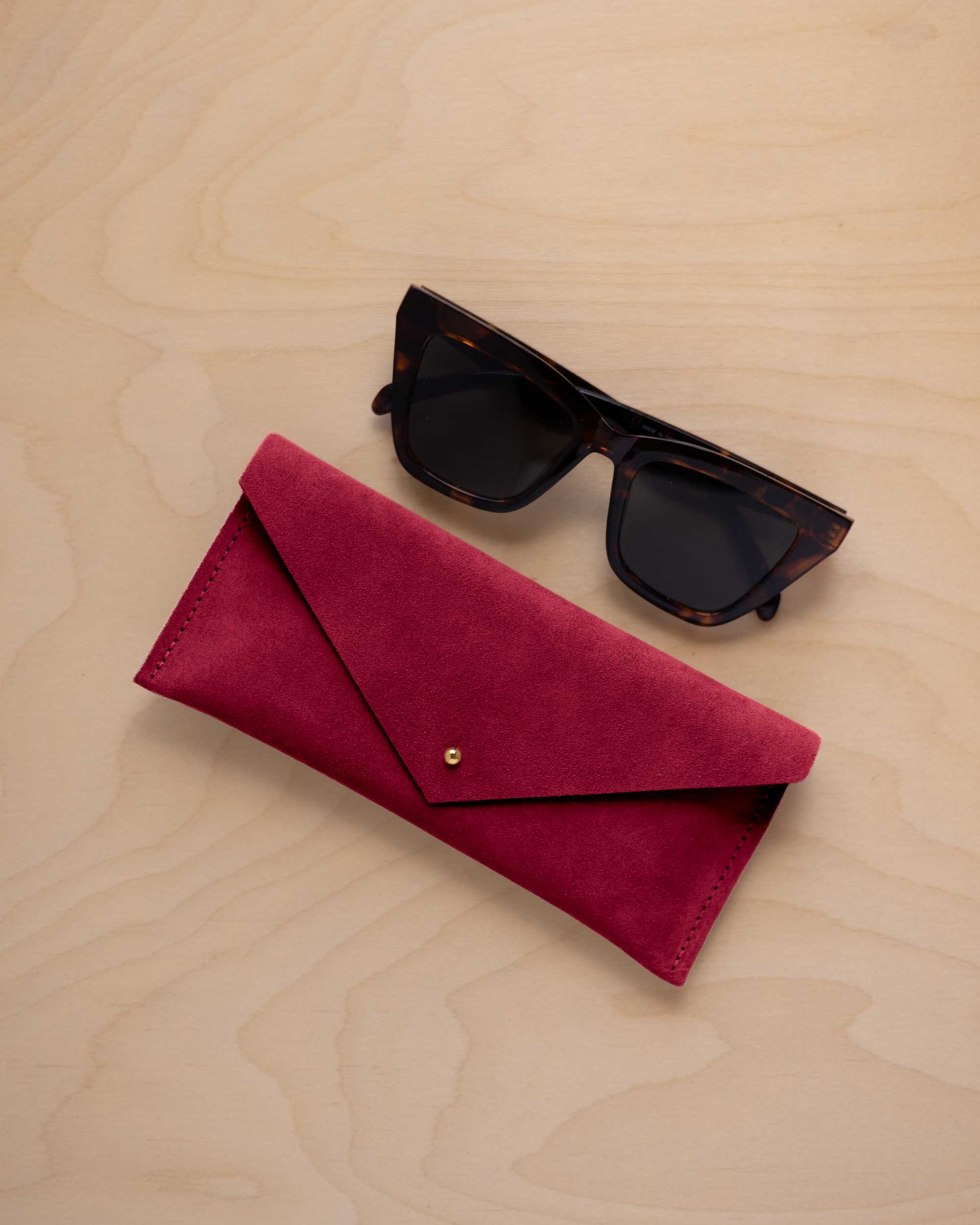 A pair of dark tortoiseshell sunglasses sit next to a crimson red suede envelope style eye glasses case with a small gold button, set on a light wood grain background.