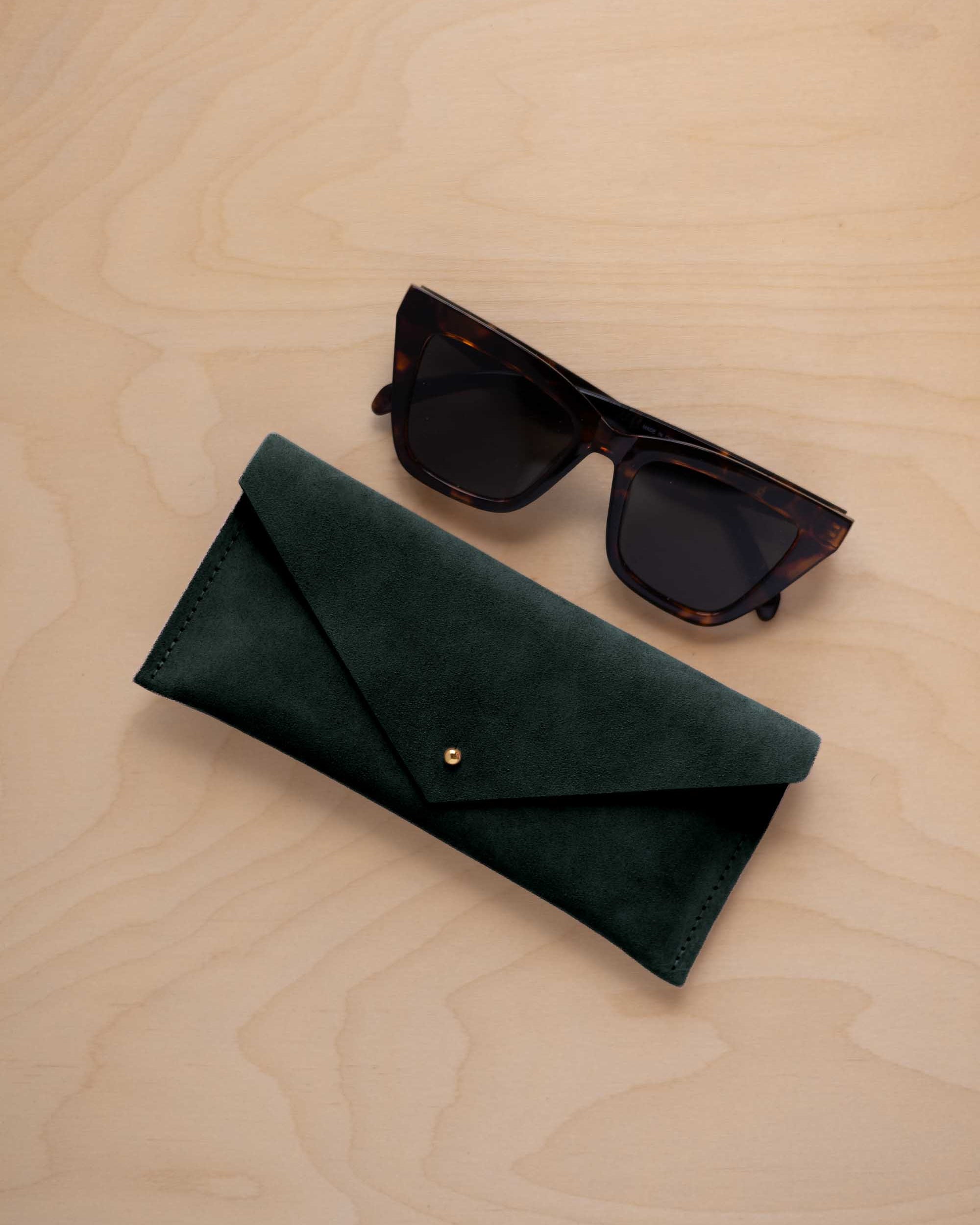 A pair of dark tortoiseshell sunglasses sit next to a deep forest green suede envelope style eye glasses case with a small gold button, set on a light wood grain background.