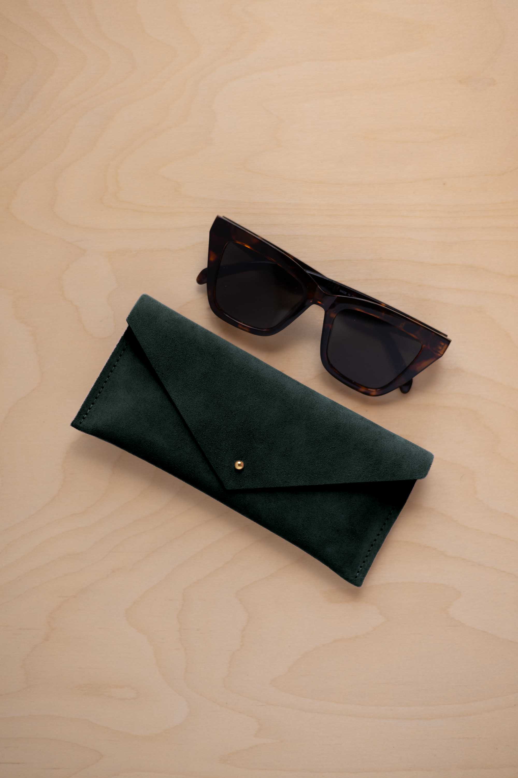 A pair of dark tortoiseshell sunglasses sit next to a deep forest green suede envelope style eye glasses case with a small gold button, set on a light wood grain background.