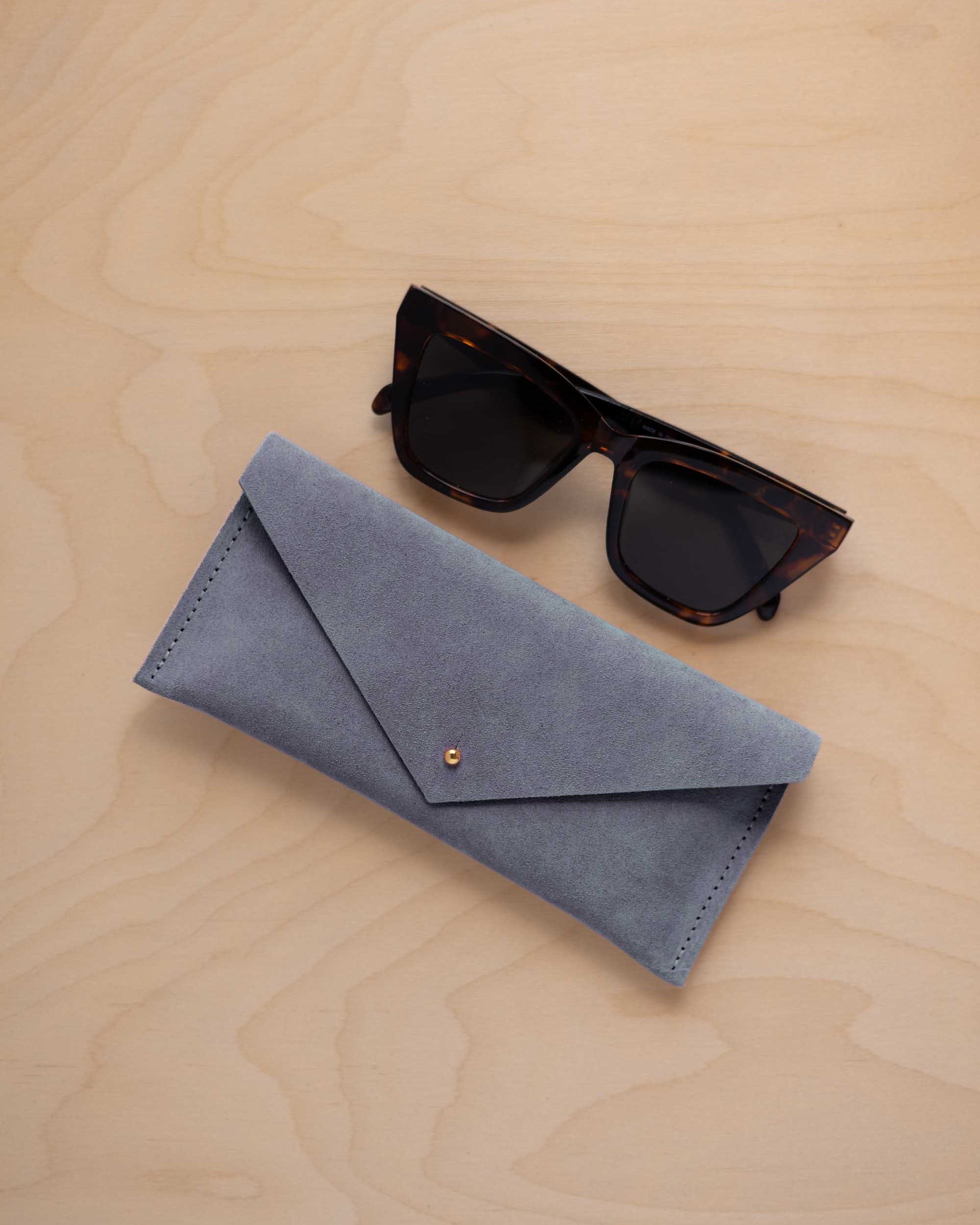 A pair of dark tortoiseshell sunglasses sit next to a dusky blue suede envelope style eye glasses case with a small gold button, set on a light wood grain background.